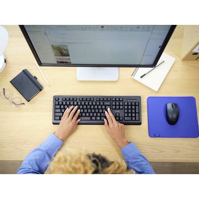Eine Person tippt auf einer schwarzen Tastatur vor einem Computerbildschirm. Auf dem Tisch liegen ein Notizbuch, eine Brille und eine Maus.