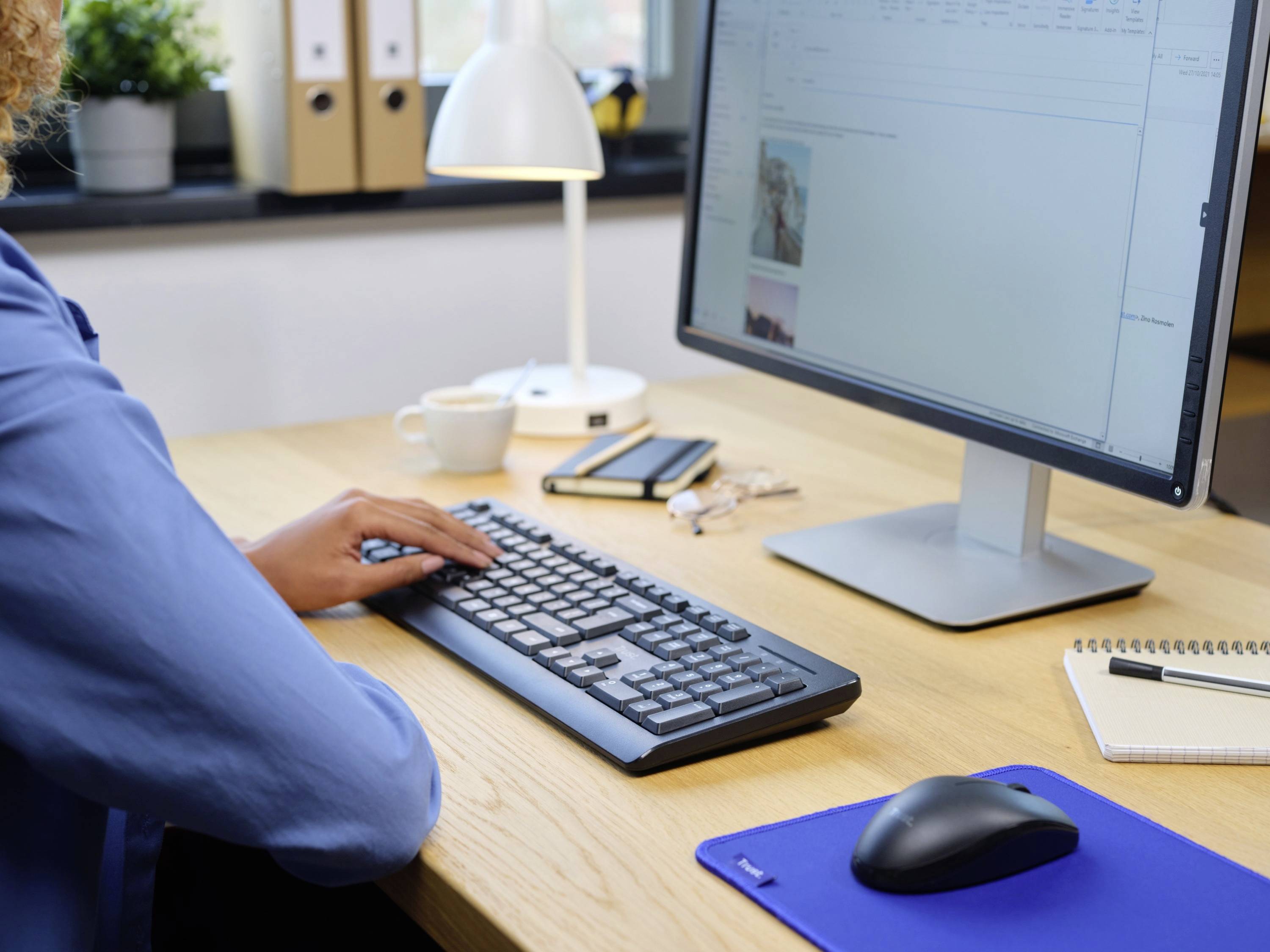 Eine Person arbeitet an einem Schreibtisch am Computer. Vorhanden sind Tastatur, Maus, Notizbuch und Tasse, Innenraumumgebung.