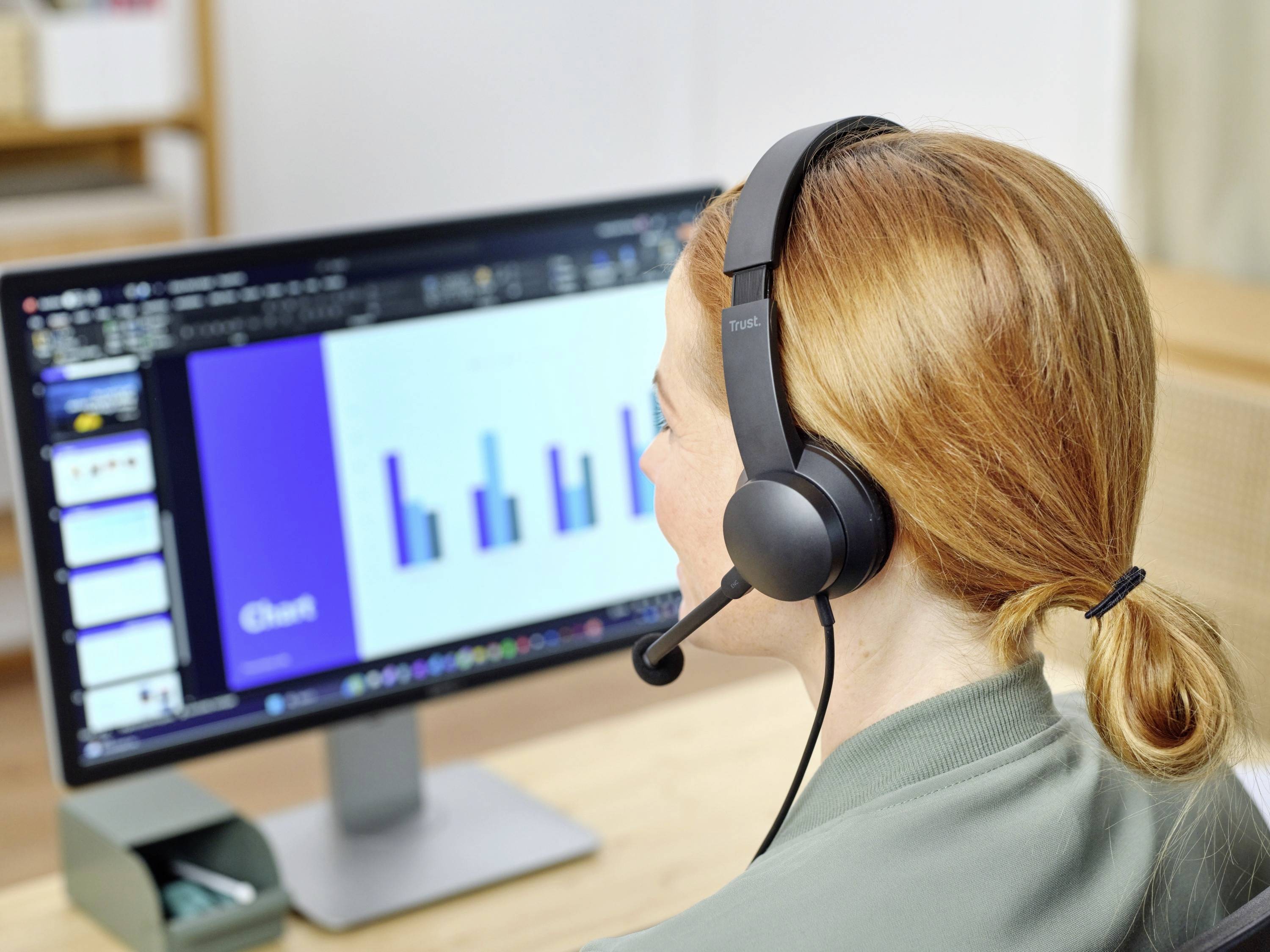Eine Frau mit Headset sitzt vor einem Computerbildschirm, der ein Balkendiagramm anzeigt. Im Hintergrund sind Büromöbel sichtbar.
