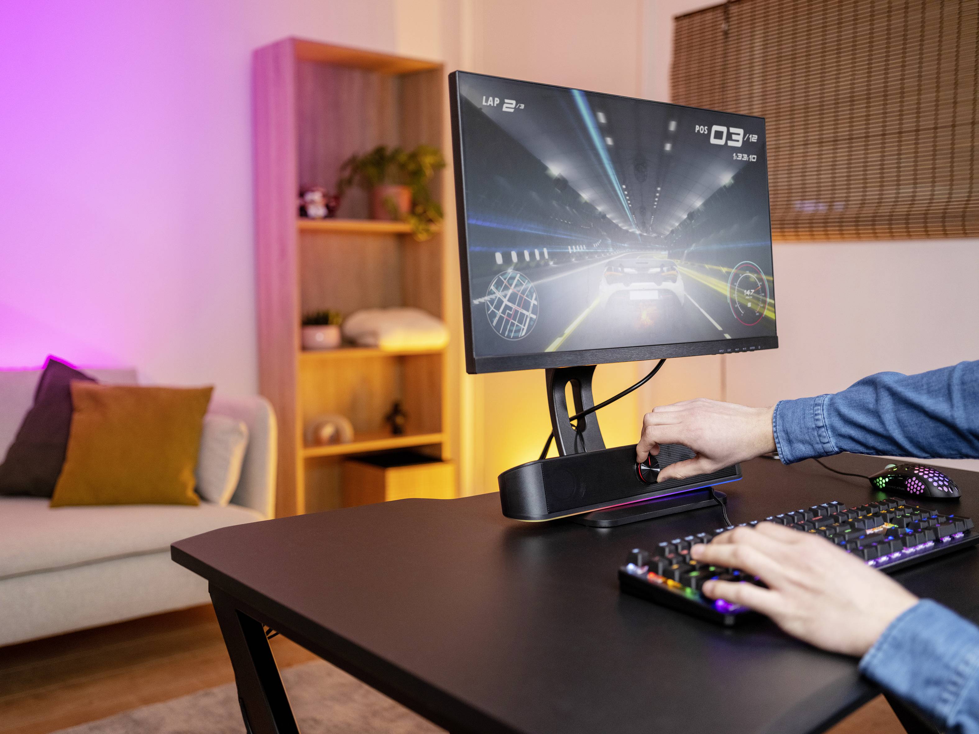 Person spielt ein Autorennspiel an einem Monitor auf einem Schreibtisch mit Tastatur und bunter Beleuchtung. Wohnzimmer im Hintergrund.
