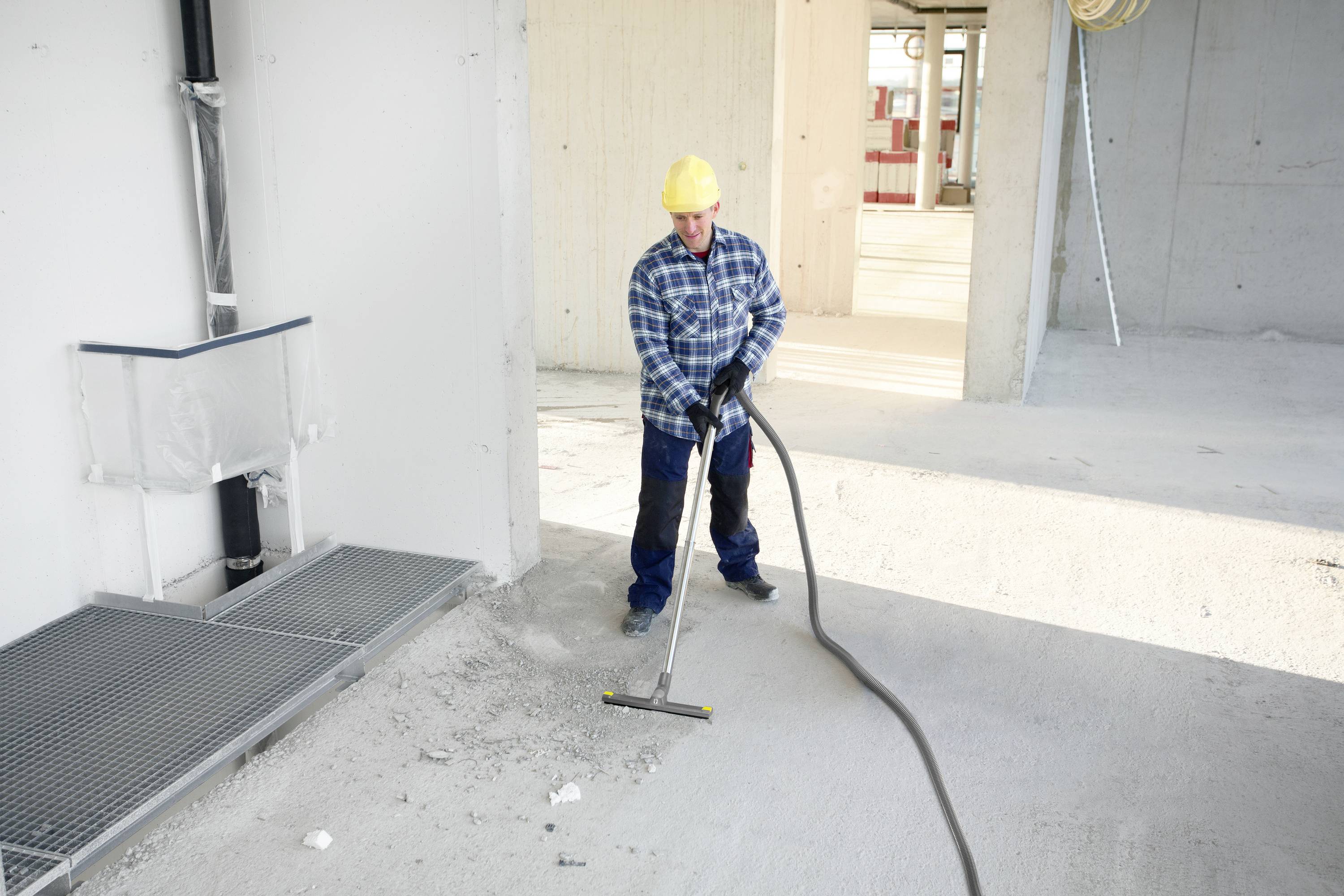 Ein Bauarbeiter trägt einen gelben Helm und blaue Arbeitskleidung. Er reinigt eine Baustelle mit einem Industriestaubsauger.