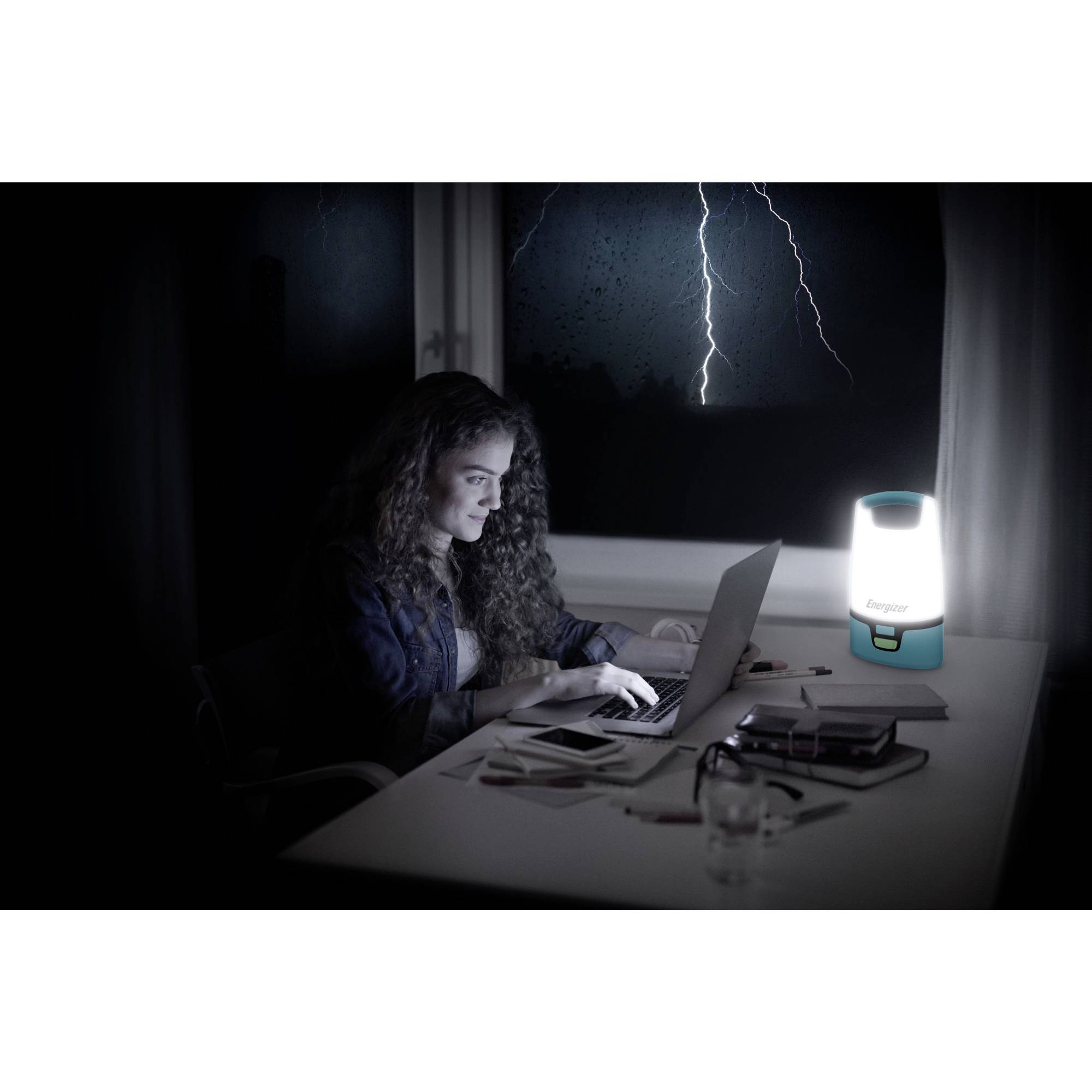 Eine Frau arbeitet nachts bei Gewitter am Laptop. Eine Lampe erhellt den Tisch. Blitze sind durch das Fenster sichtbar.