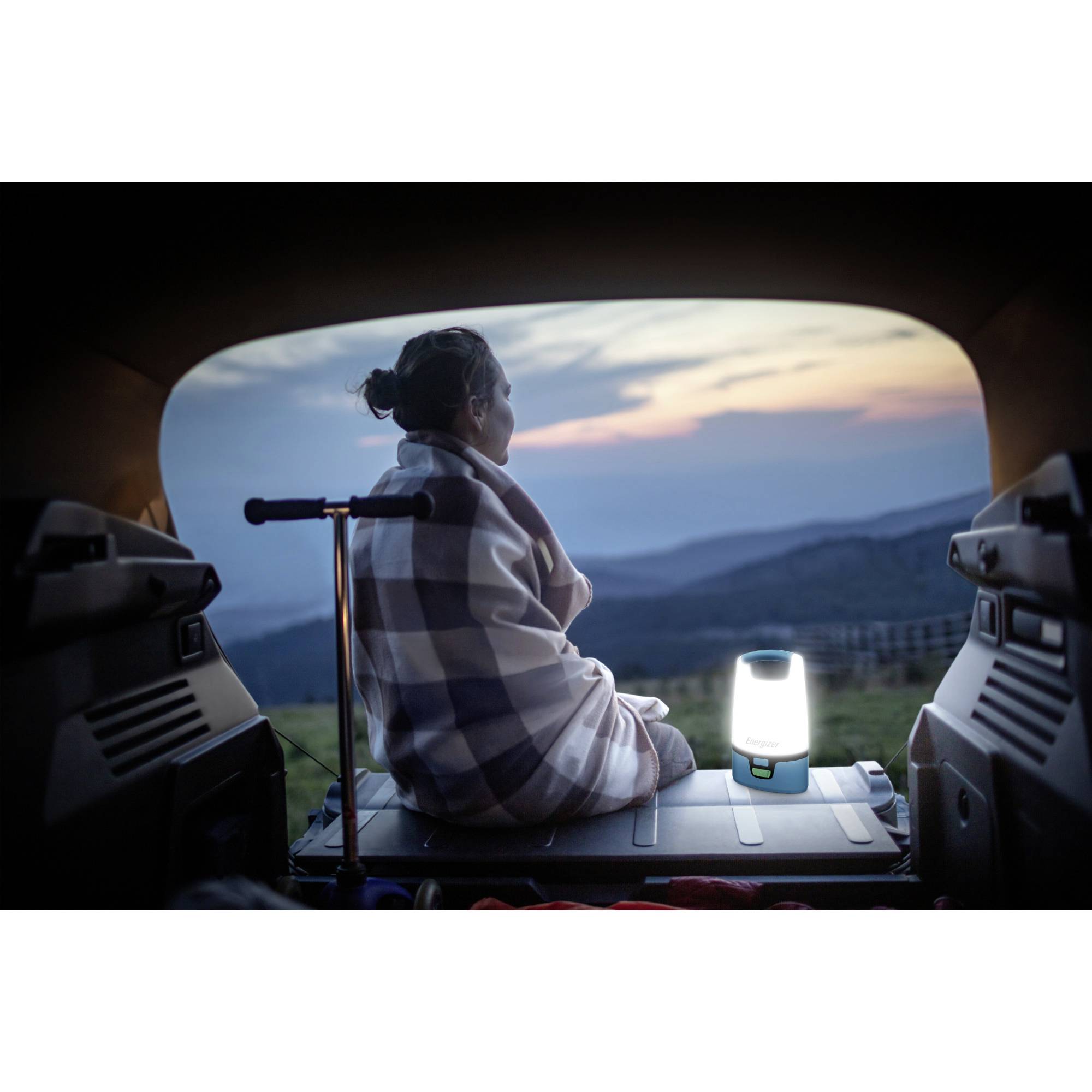 Eine Person sitzt in einem Auto, eingewickelt in eine Decke, blickt auf eine ruhige Landschaft im Abendlicht mit einer leuchtenden Lampe daneben.