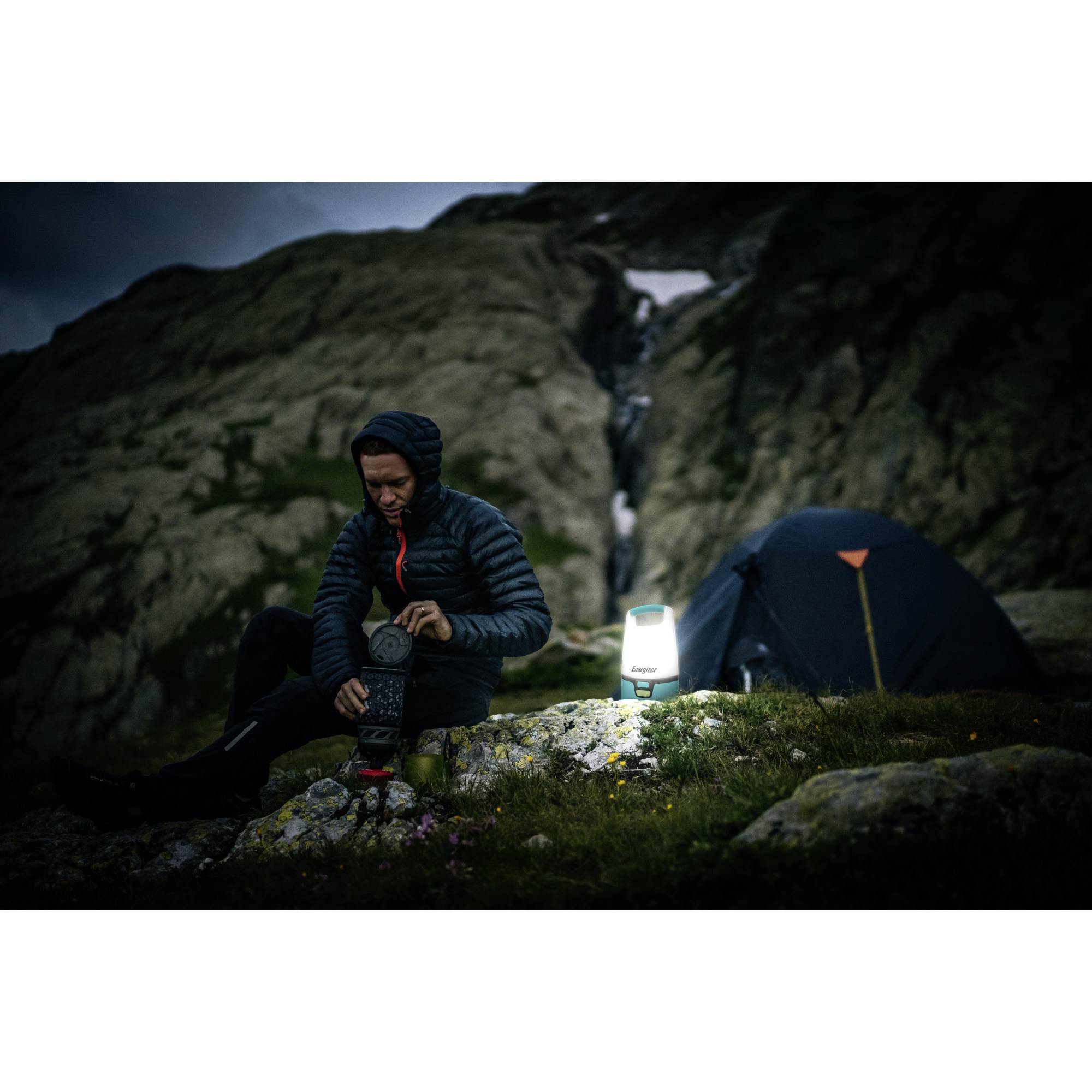 Eine Person sitzt im Freien vor einem Zelt in einer bergigen Landschaft und bereitet eine Mahlzeit mit einem tragbaren Kocher zu. Es ist Abenddämmerung.