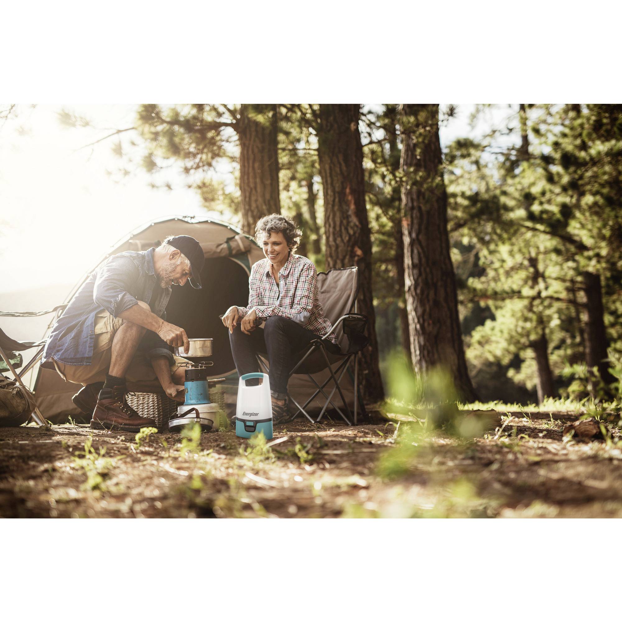 Ein älteres Paar sitzt vor einem Zelt im Wald. Der Mann kocht mit einem Campingkocher, während die Frau daneben sitzt und zuschaut.