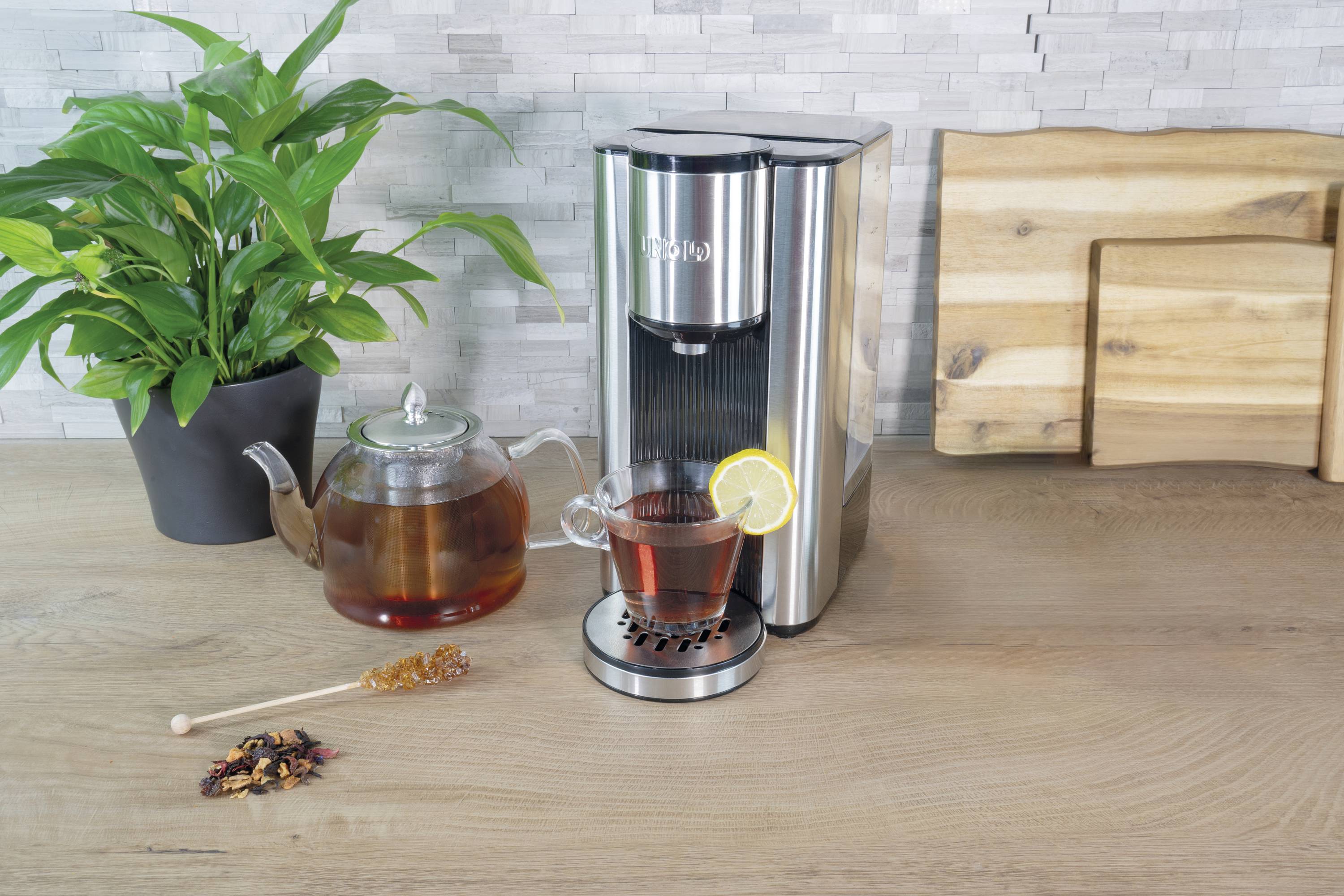 Eine Teemaschine auf dem Tisch mit einer Tasse Tee und Teekanne, daneben eine Pflanze und Holzbrett, in einer Küche platziert.