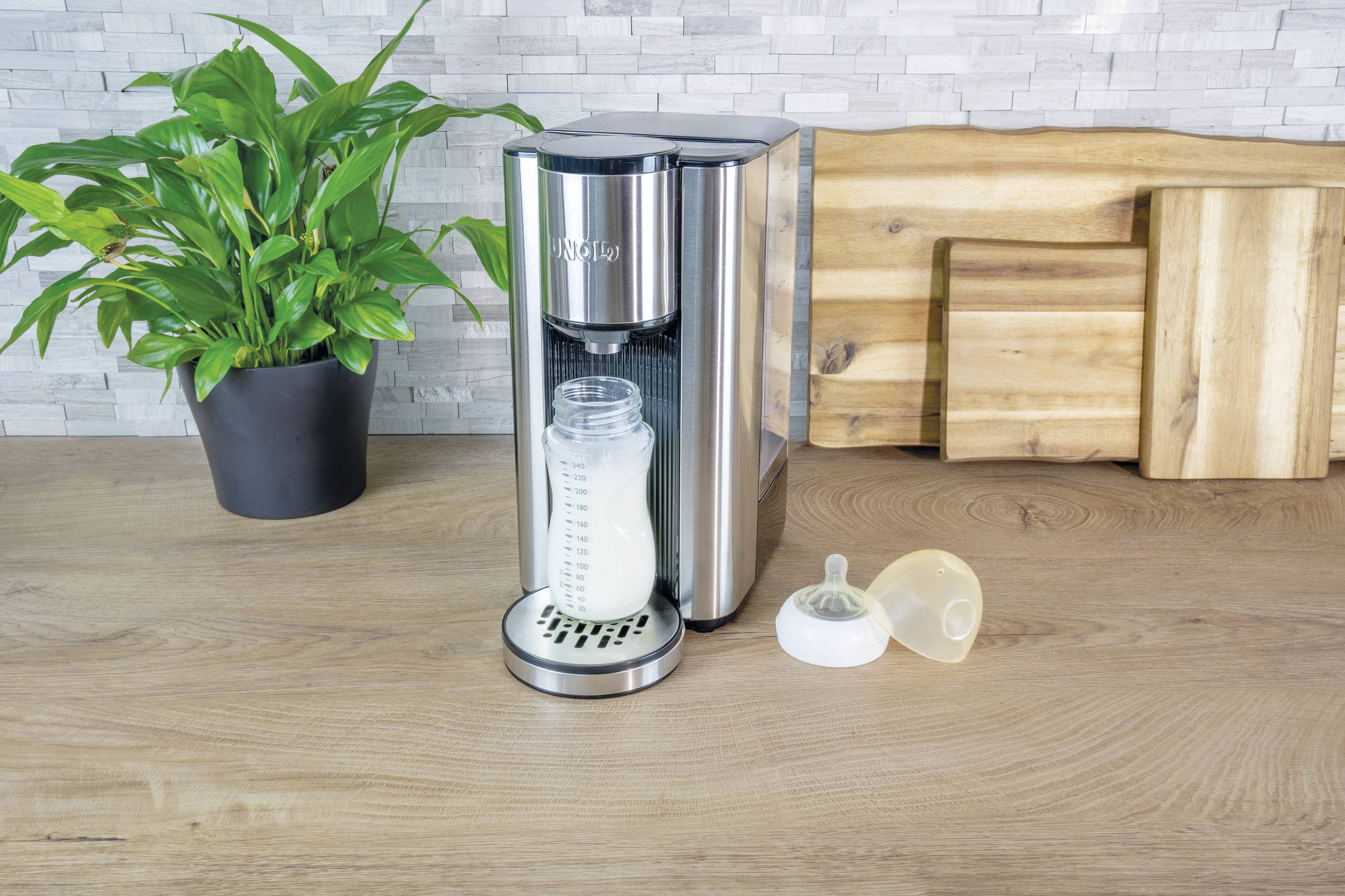 Ein Milchzubereitungsgerät auf einer Küchenarbeitsplatte, neben einem mit Milch gefüllten Fläschchen und einem offenen Flaschendeckel.