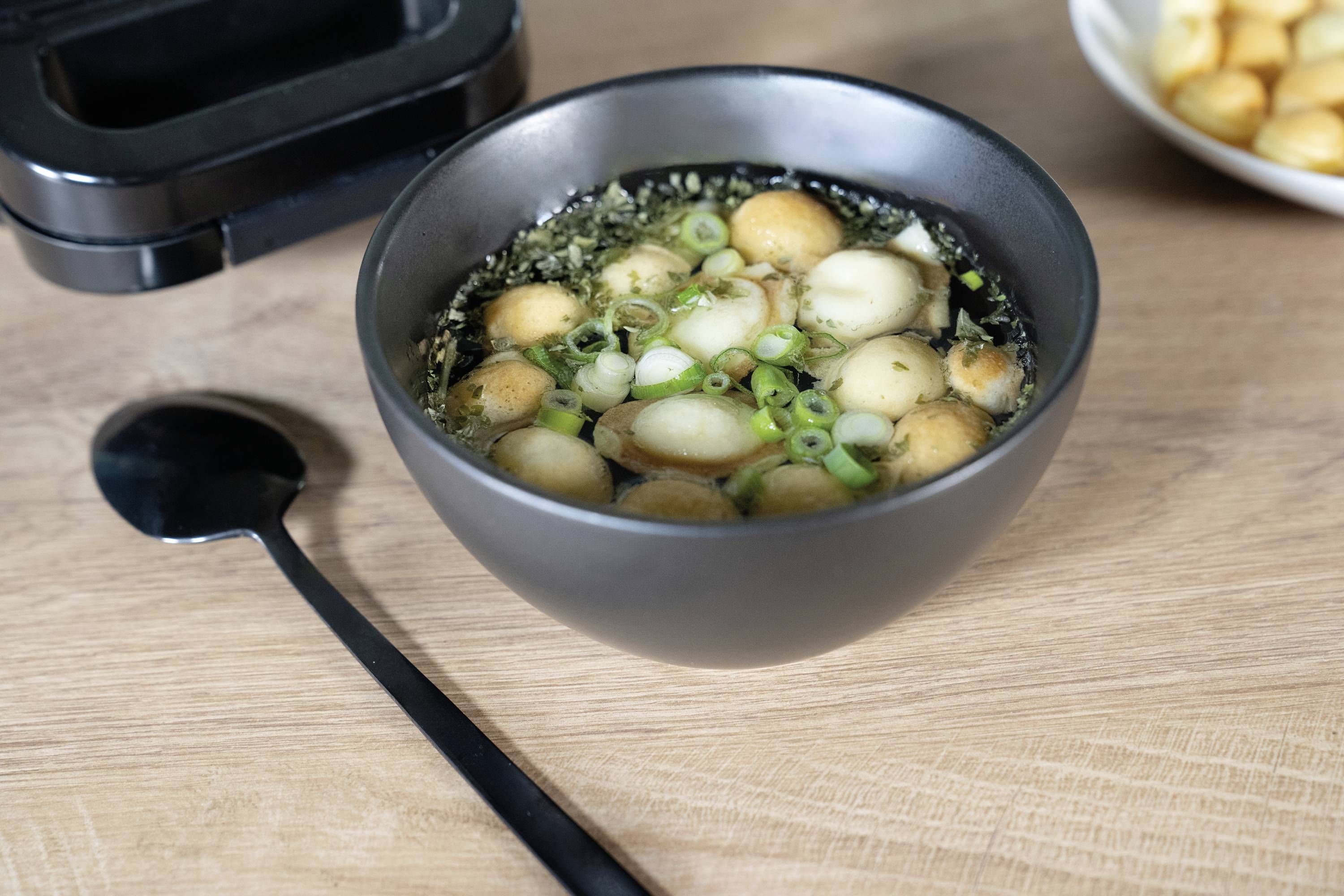 Eine schwarze Schüssel mit klarer Suppe, darin schwimmende Brotcroutons, grüne Zwiebeln und Kräuter. Daneben liegt ein schwarzer Löffel.