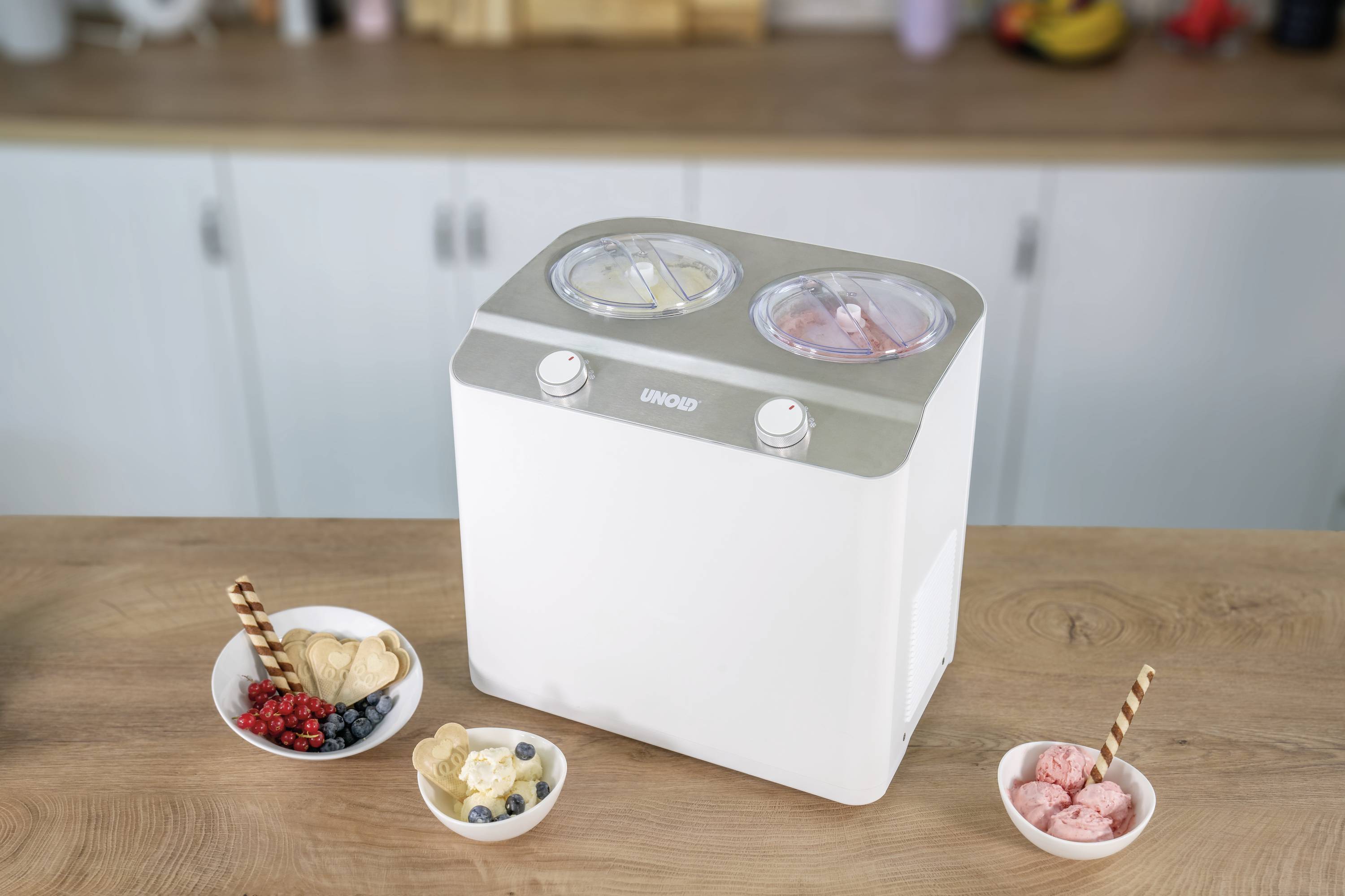 Eiscreme-Maschine mit zwei Schüsseln, gefüllt mit gelber und rosafarbener Eiscreme. Umgeben von Schalen mit verschiedenen Toppings.