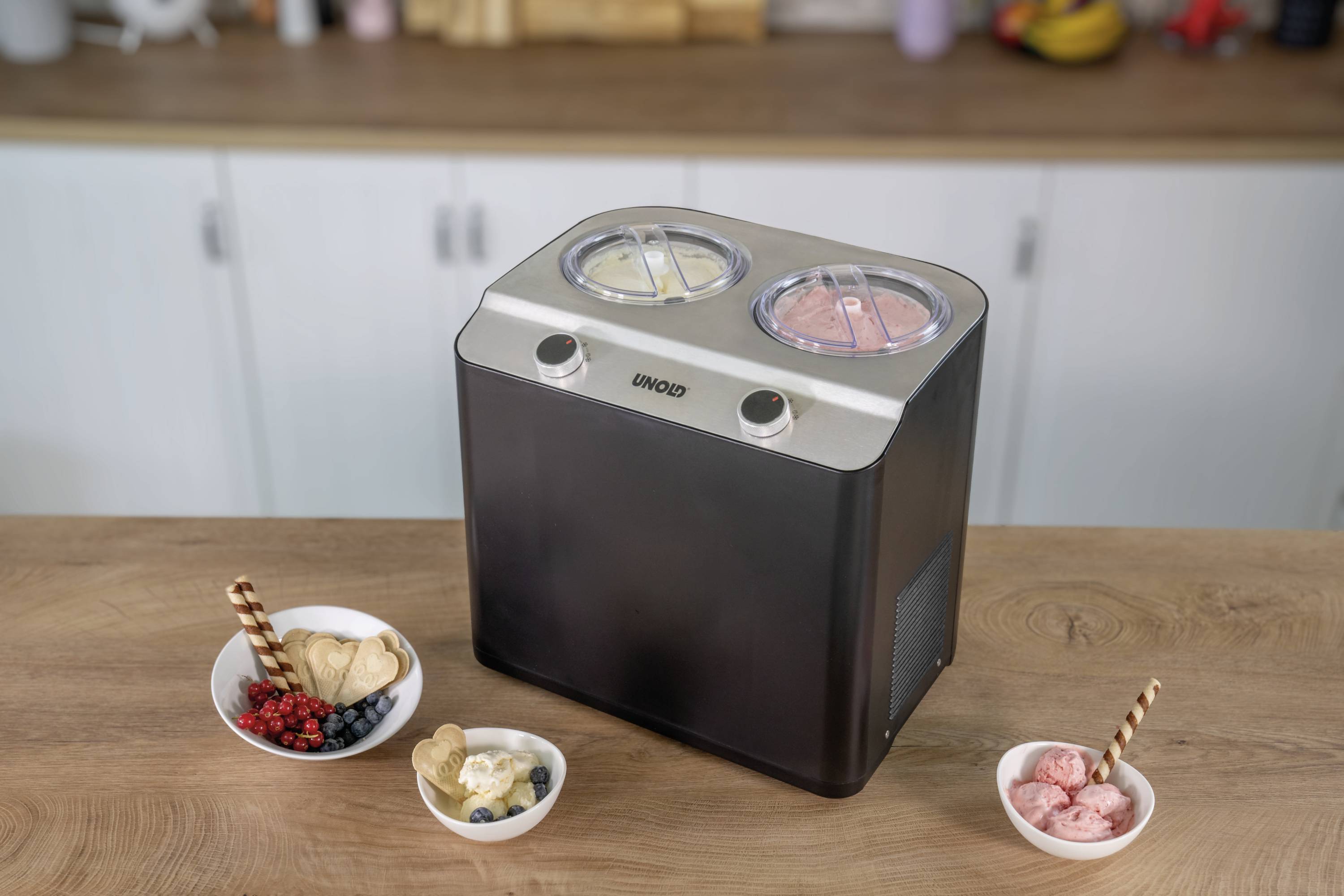 Eismaschine mit zwei Behältern, gefüllt mit Vanille- und Erdbeereis. Drei Schüsseln mit Eis und Früchten stehen davor auf einem Tisch.