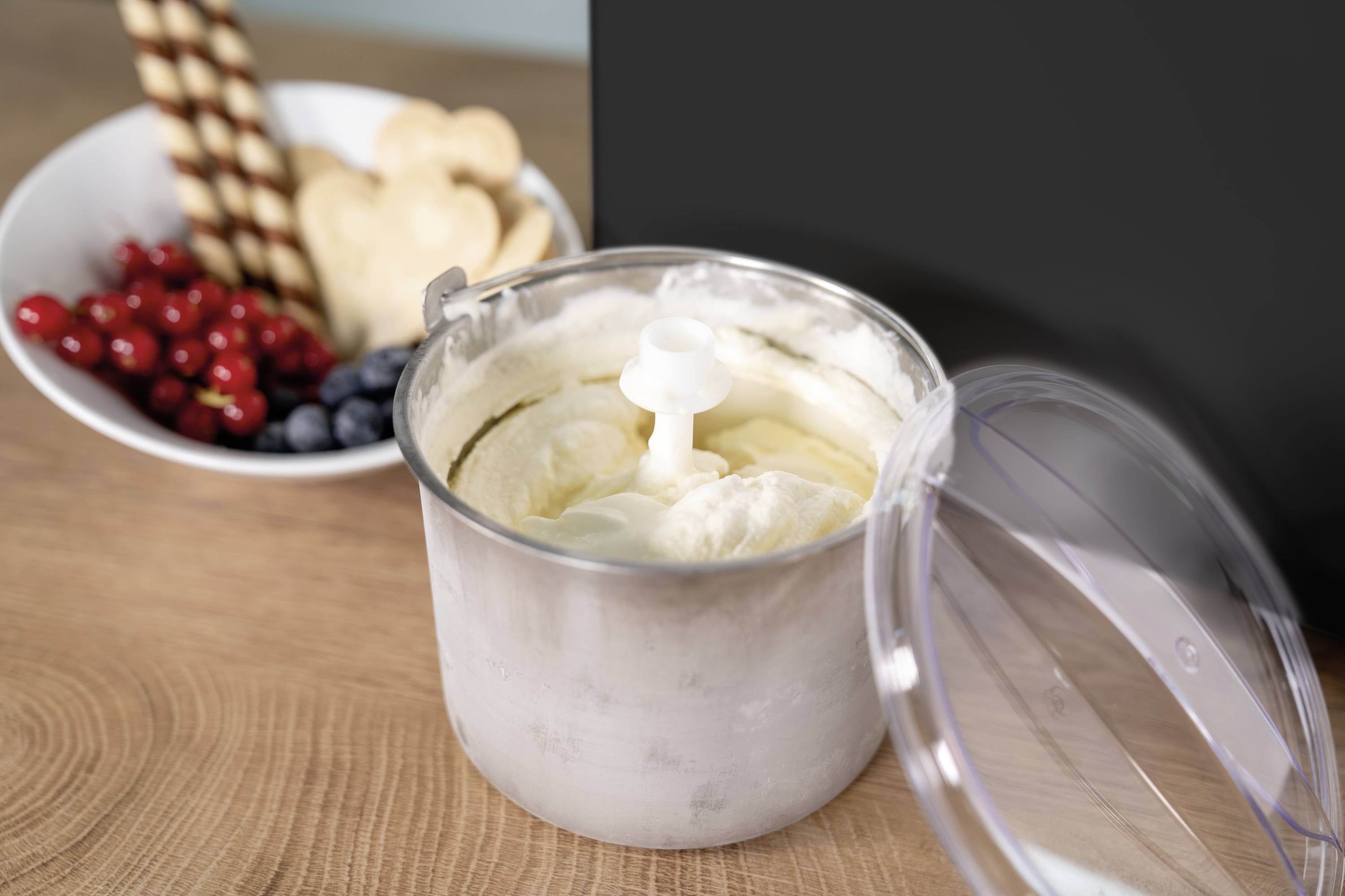 Eiscreme in einer Metall-Schüssel mit Deckel auf Holzoberfläche; im Hintergrund stehen Kekse und Früchte in einer weißen Schale.