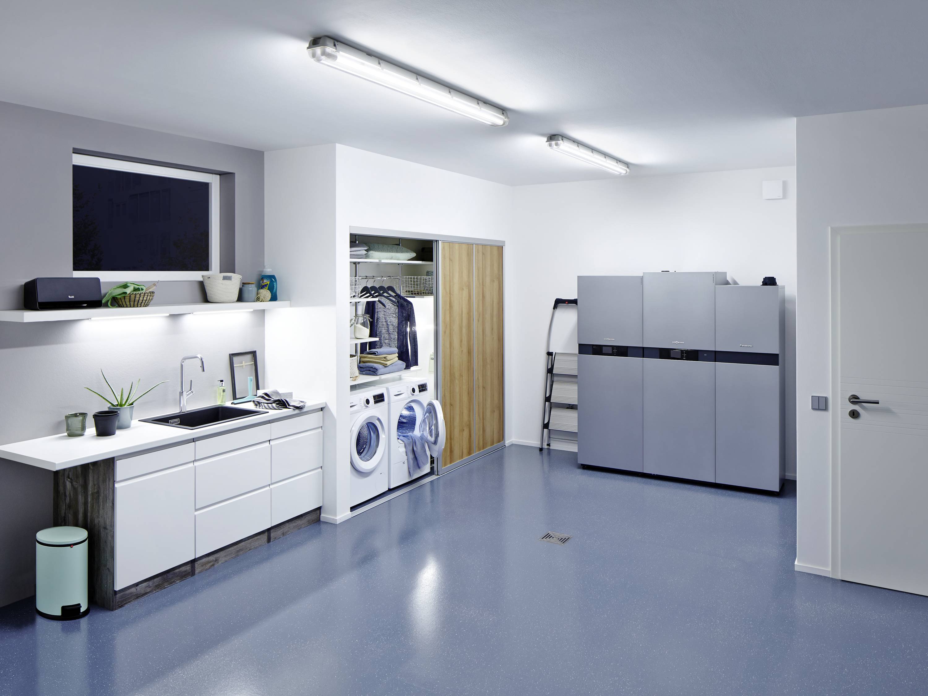 Ein moderner Hauswirtschaftsraum mit Waschmaschine, Trockner und Stauraum. Links sind eine Spüle und Regale mit Pflanzen.