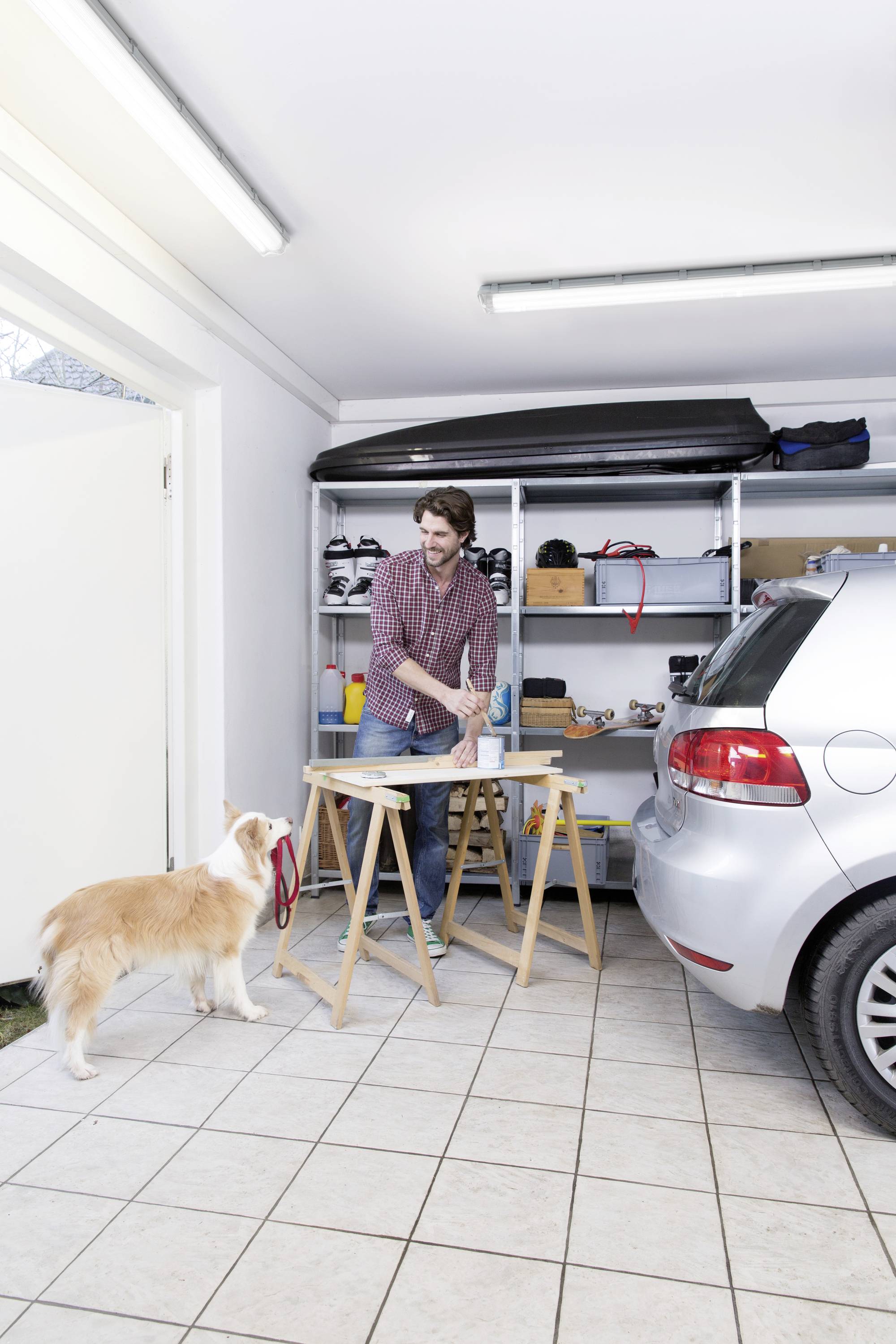 Ein Mann arbeitet an einem Tisch in einer Garage, während ein Hund ihm aufmerksam zusieht. Links ist die Tür geöffnet, rechts steht ein Auto.