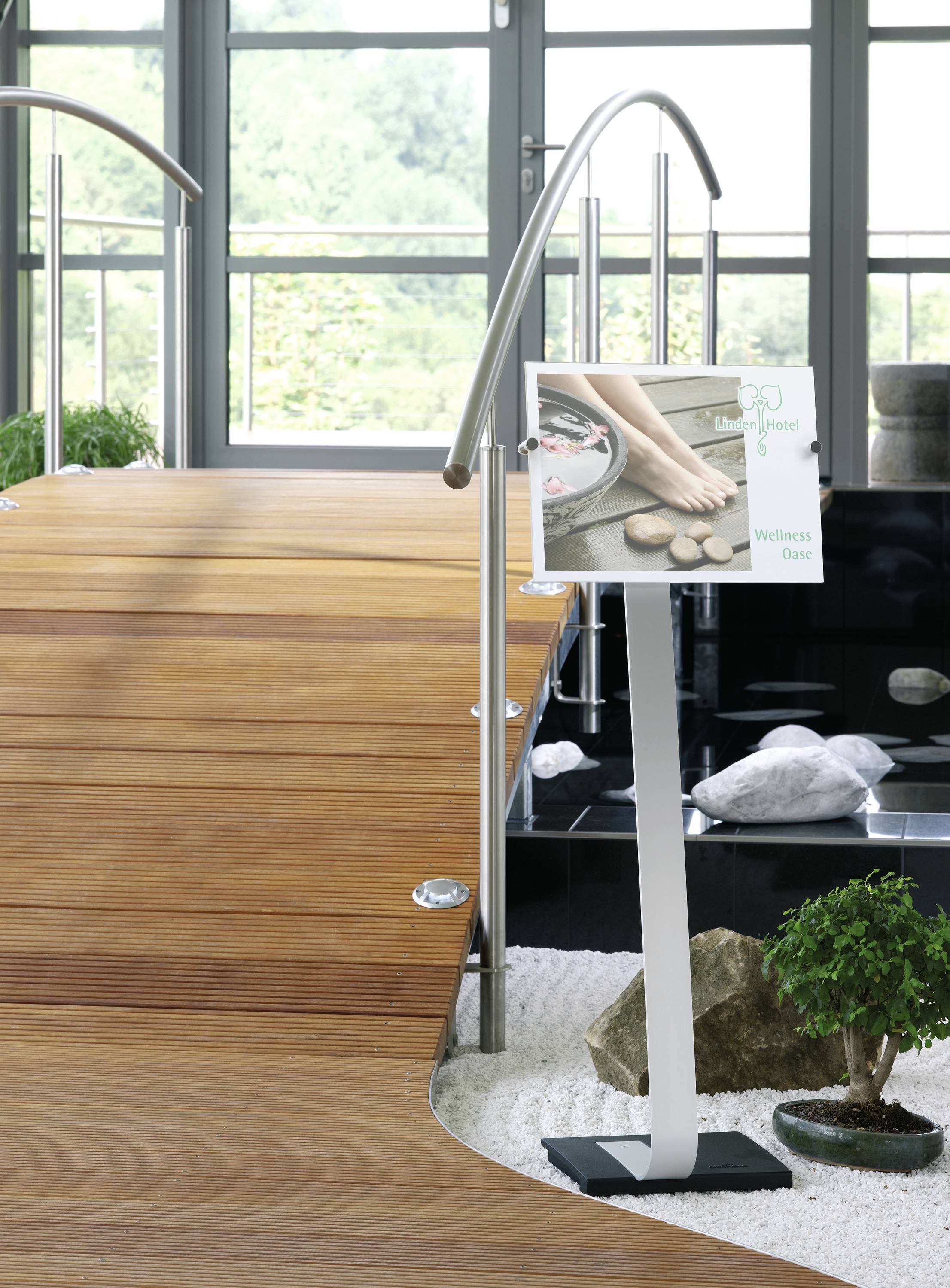 Ein Holzsteg mit Geländer führt durch einen Raum mit großen Fenstern. Rechts steht ein Schild mit einem Bild von Füßen auf Steinen, daneben ist ein kleiner Garten mit Pflanzen und Steinen.