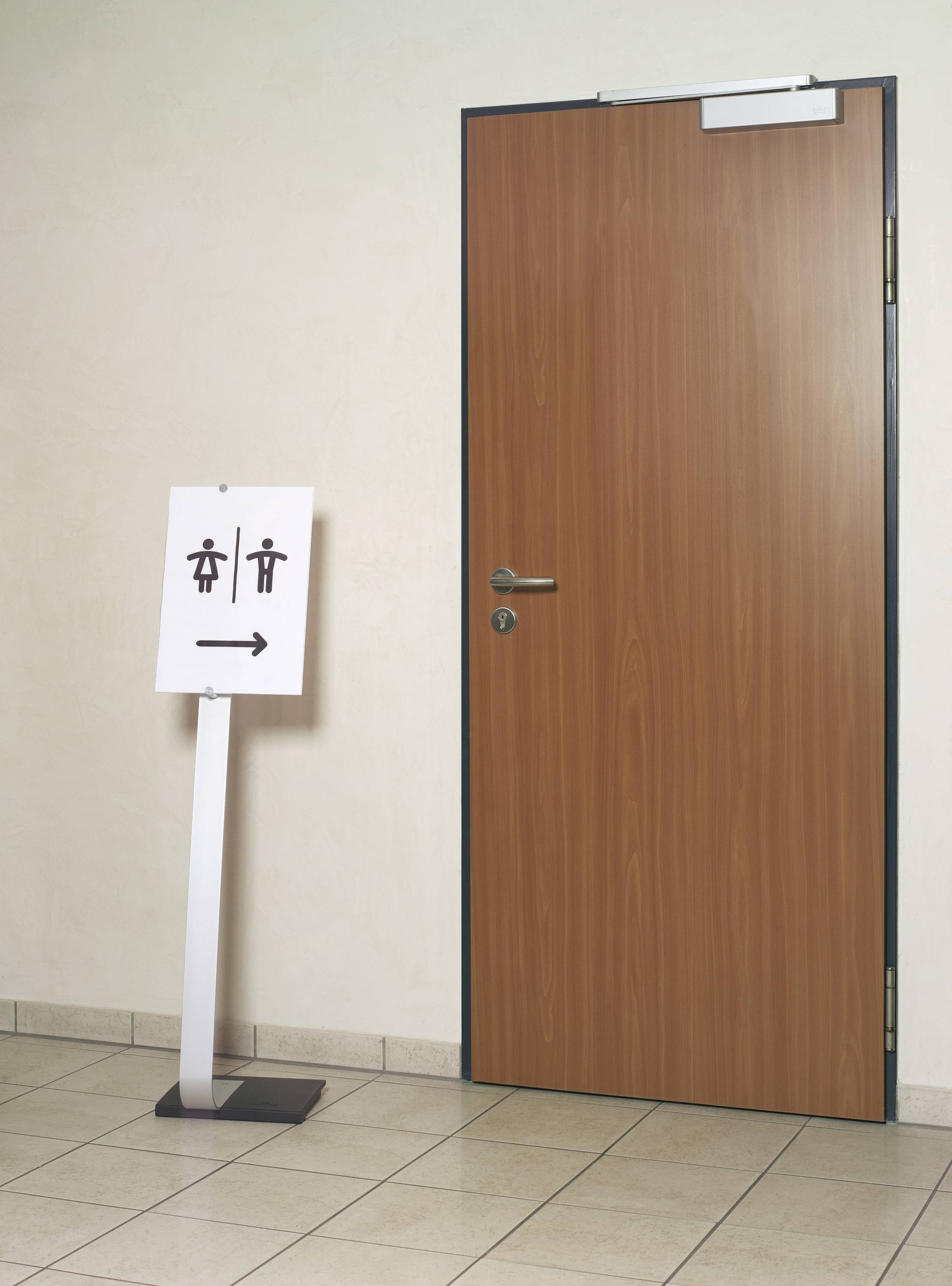 Holztür mit einem Schild davor, das auf eine Toilette hinweist. Schild zeigt Piktogramme für Männer und Frauen mit einem Pfeil nach links.