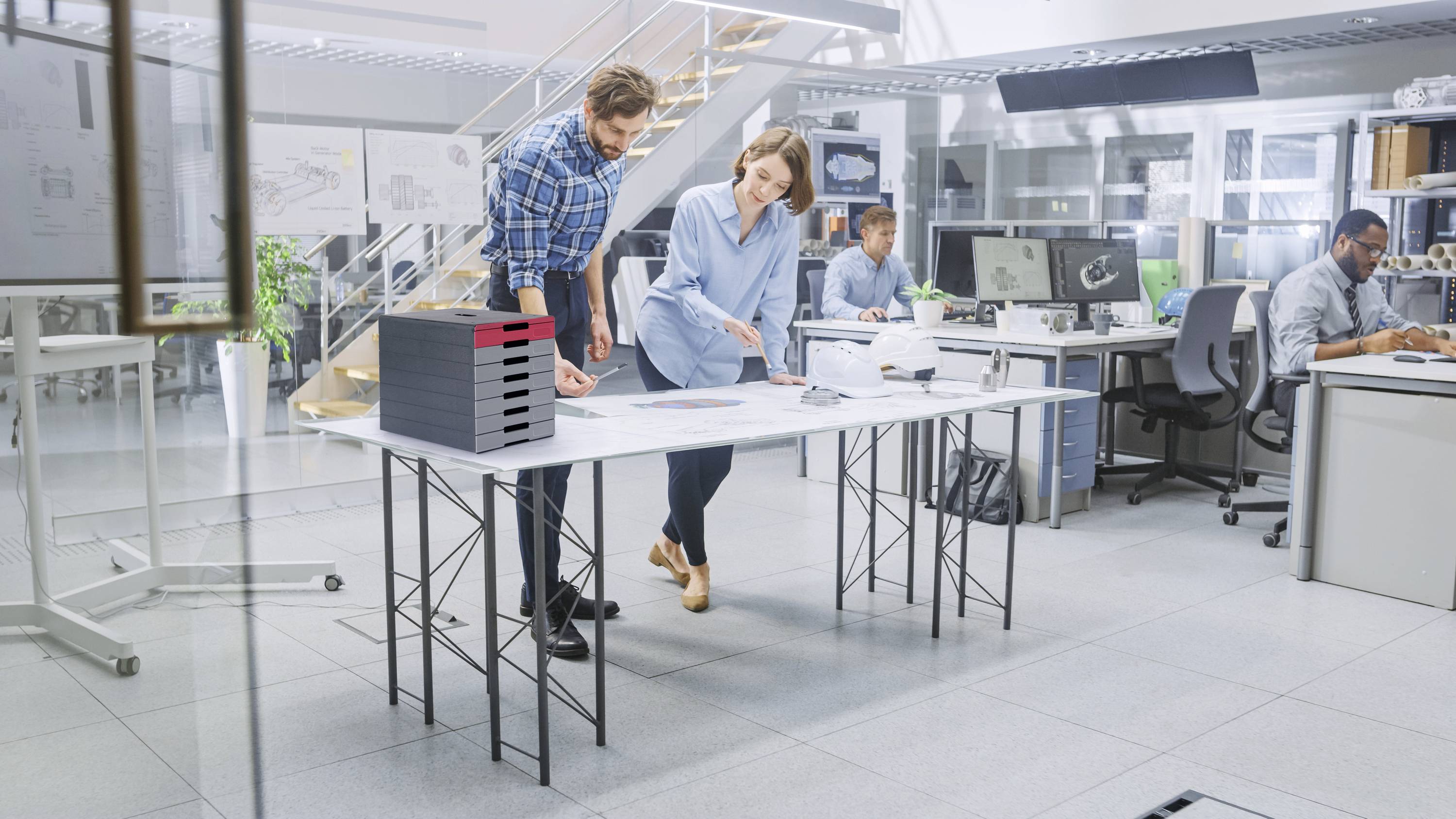 Ein Mann und eine Frau diskutieren über Dokumente auf einem Tisch in einem modernen Büro; im Hintergrund arbeiten weitere Personen.