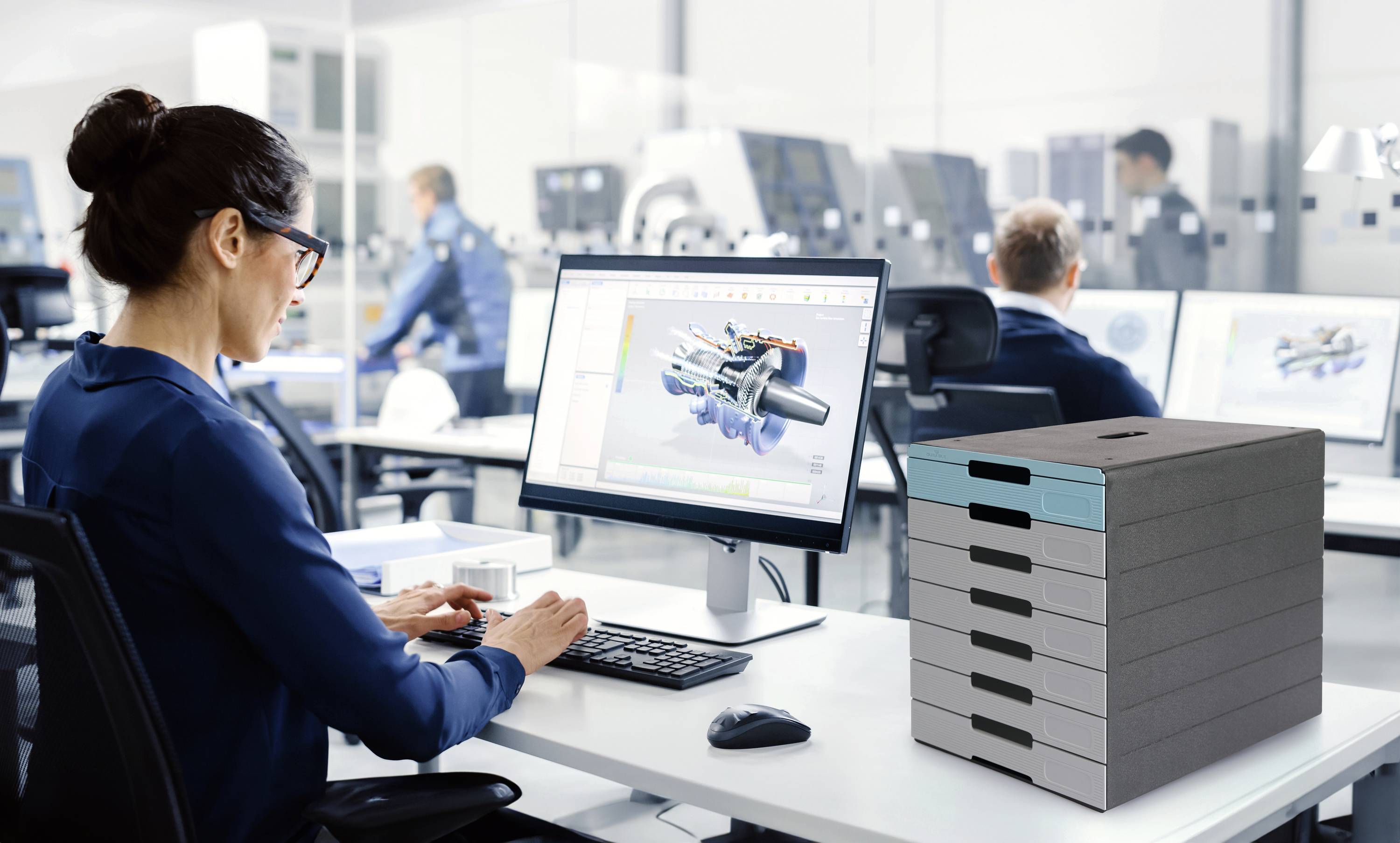 Frau arbeitet an einem Computer bei einem technischen Designprojekt in einem modernen Büro. Im Hintergrund sind Kollegen und Maschinen.