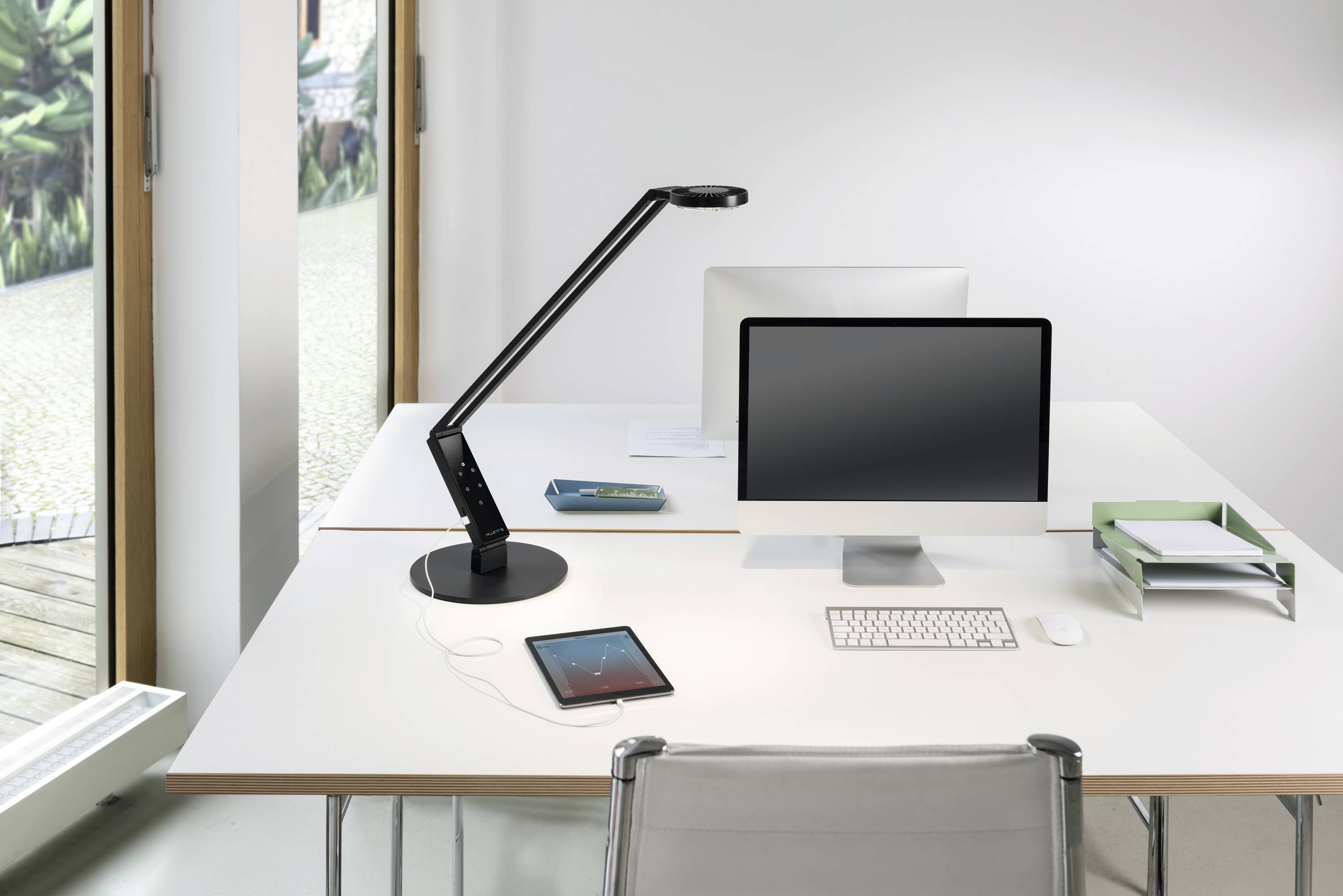 Ein aufgeräumter Schreibtisch mit einem Monitor, einer Tischlampe, einem Tablet, einem Buch und einem Papierablagefach neben einem Fenster.