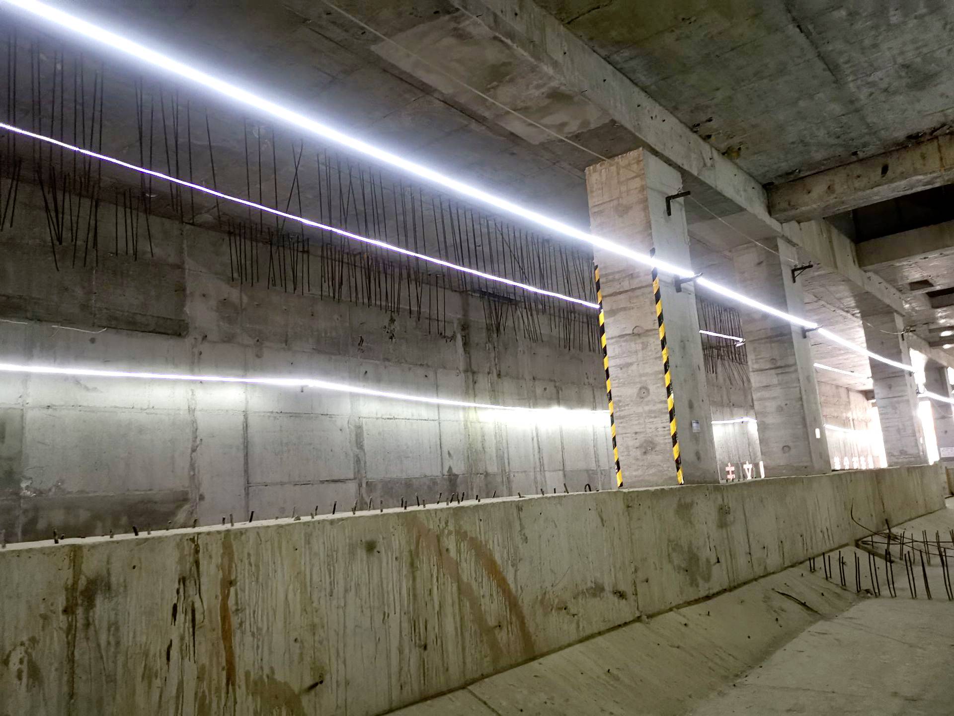 Eine Beton-Baustelle mit freiliegenden Stahlstreben und langen, leuchtenden LED-Leisten an der Decke. Die Umgebung ist weitgehend roh und unvollendet.
