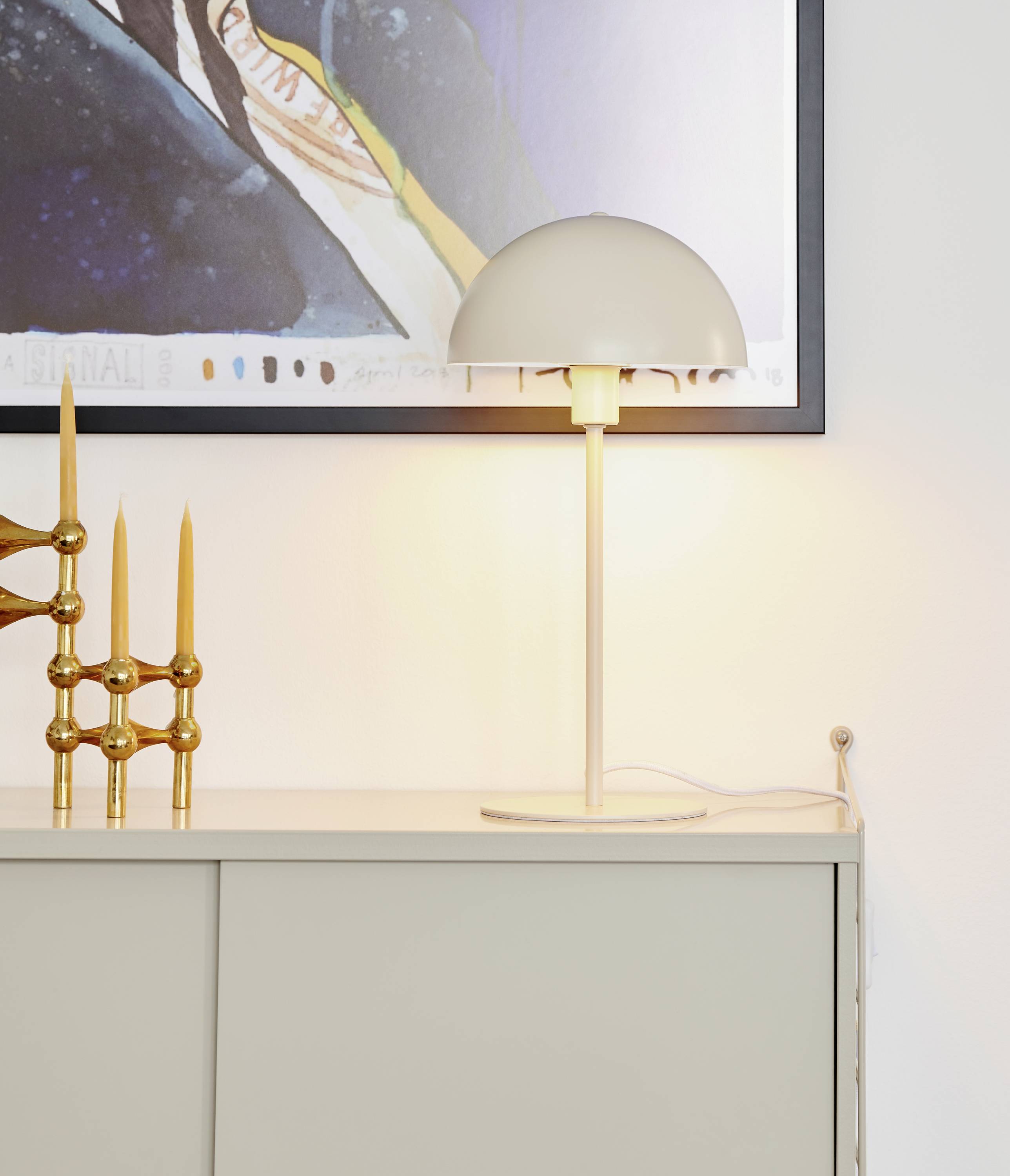 Eine Tischlampe mit rundem Schirm leuchtet auf einem weißen Schrank. Daneben steht ein dekorativer Kerzenhalter mit mehreren Kerzen.