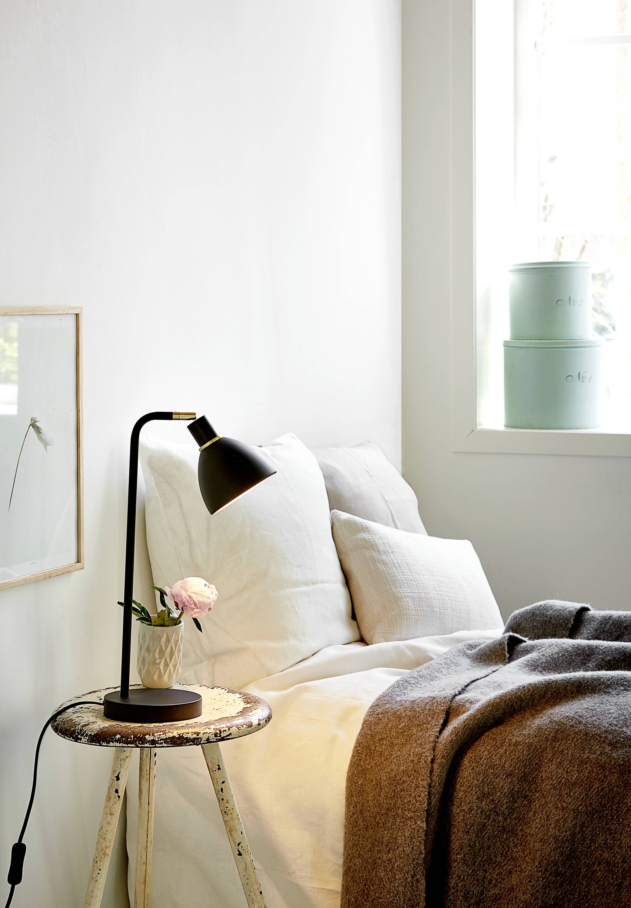 Schlafzimmer mit weißem Bett, grauer Decke, Nachttisch mit Lampe und Blume. Helle, gemütliche Raumgestaltung mit Fensterlicht.