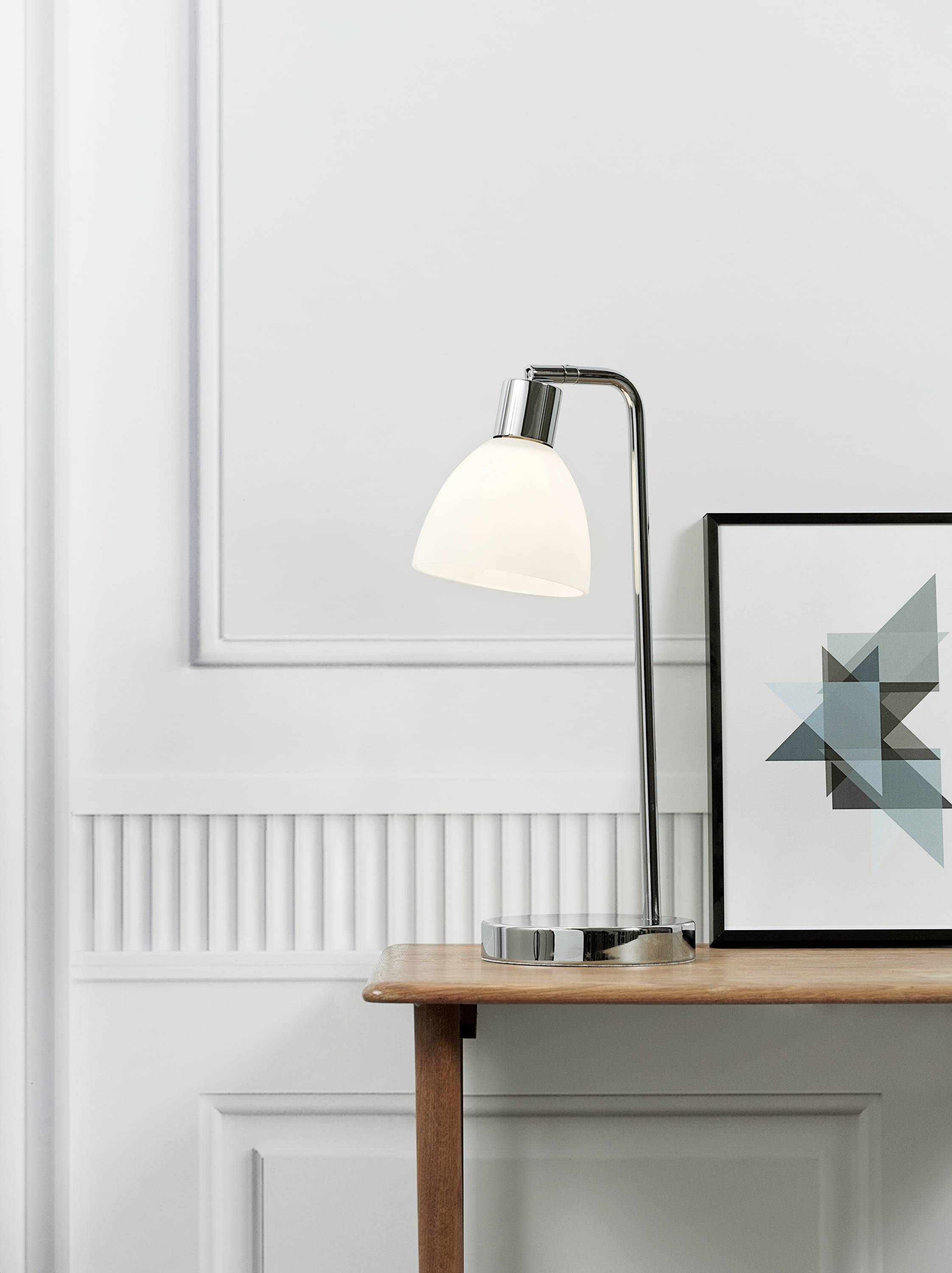 Tischlampe mit modernem Design auf einem Holztisch neben einem geometrischen Kunstbild an einer hellgrauen Wand.