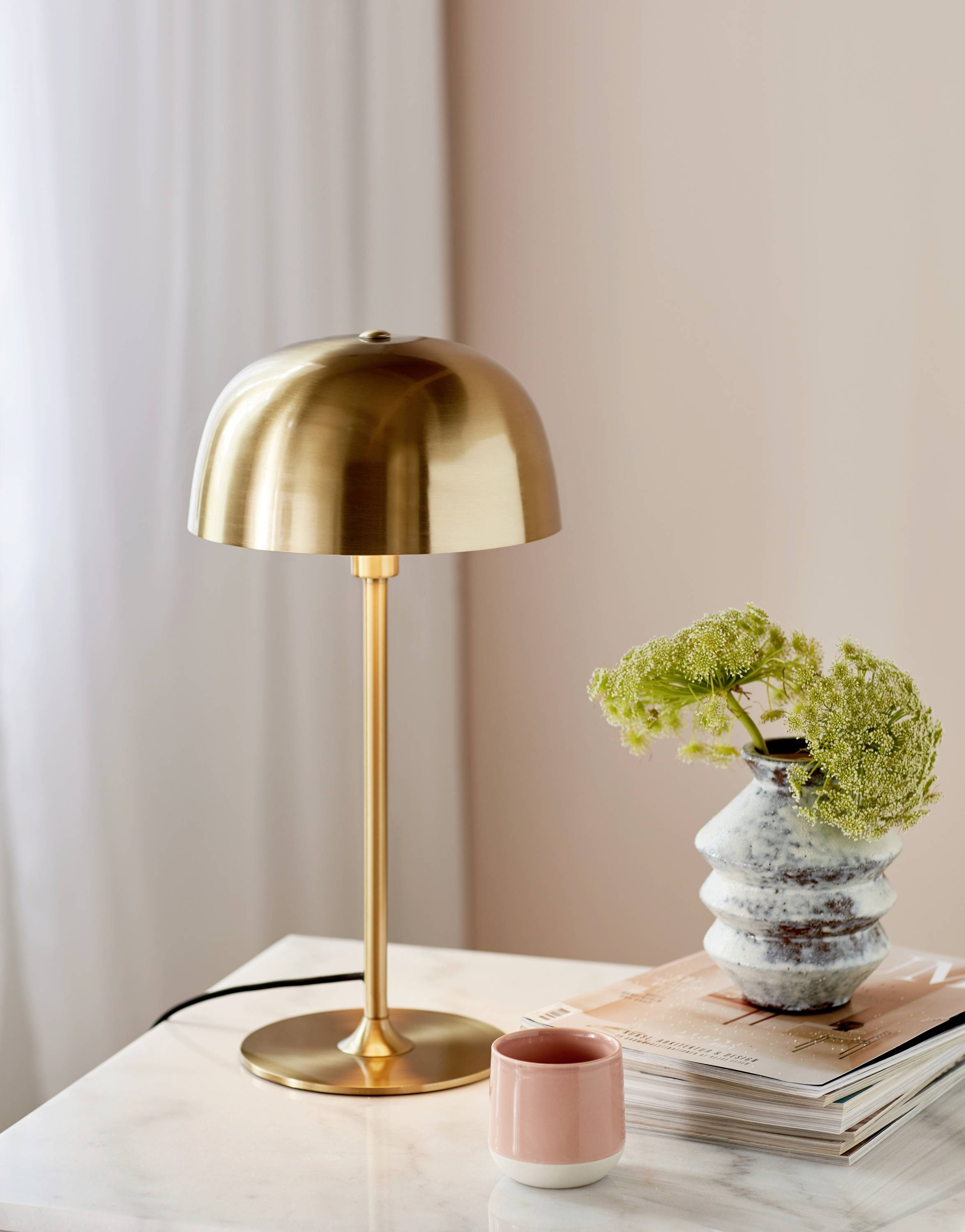Eine goldene Tischlampe auf einem Marmortisch, daneben eine Vase mit Grünpflanze und ein rosa Becher auf Büchern. Helle, gemütliche Umgebung.