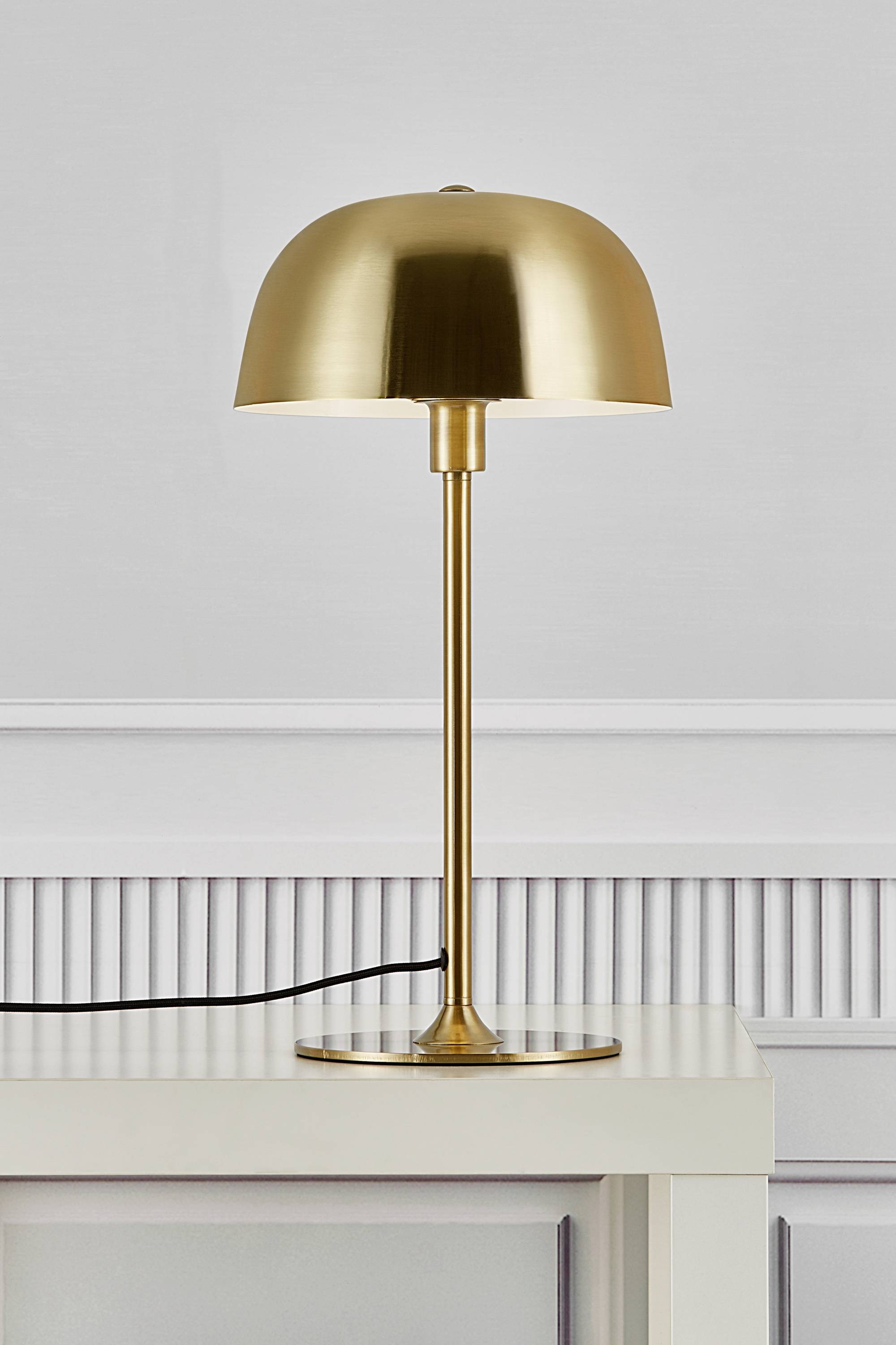 Eine goldene Tischlampe mit rundem Schirm steht auf einem weißen Tisch vor einer grauen Wand.