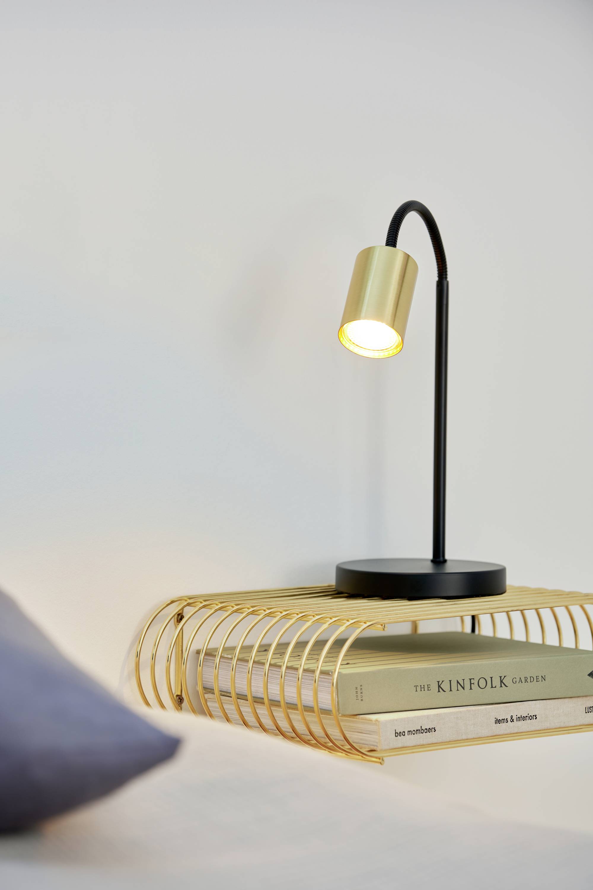 Eine elegante Nachttischlampe aus Metall leuchtet sanft auf Bücher, darunter 'The Kinfolk Garden', auf einem goldenen Gitterregal.