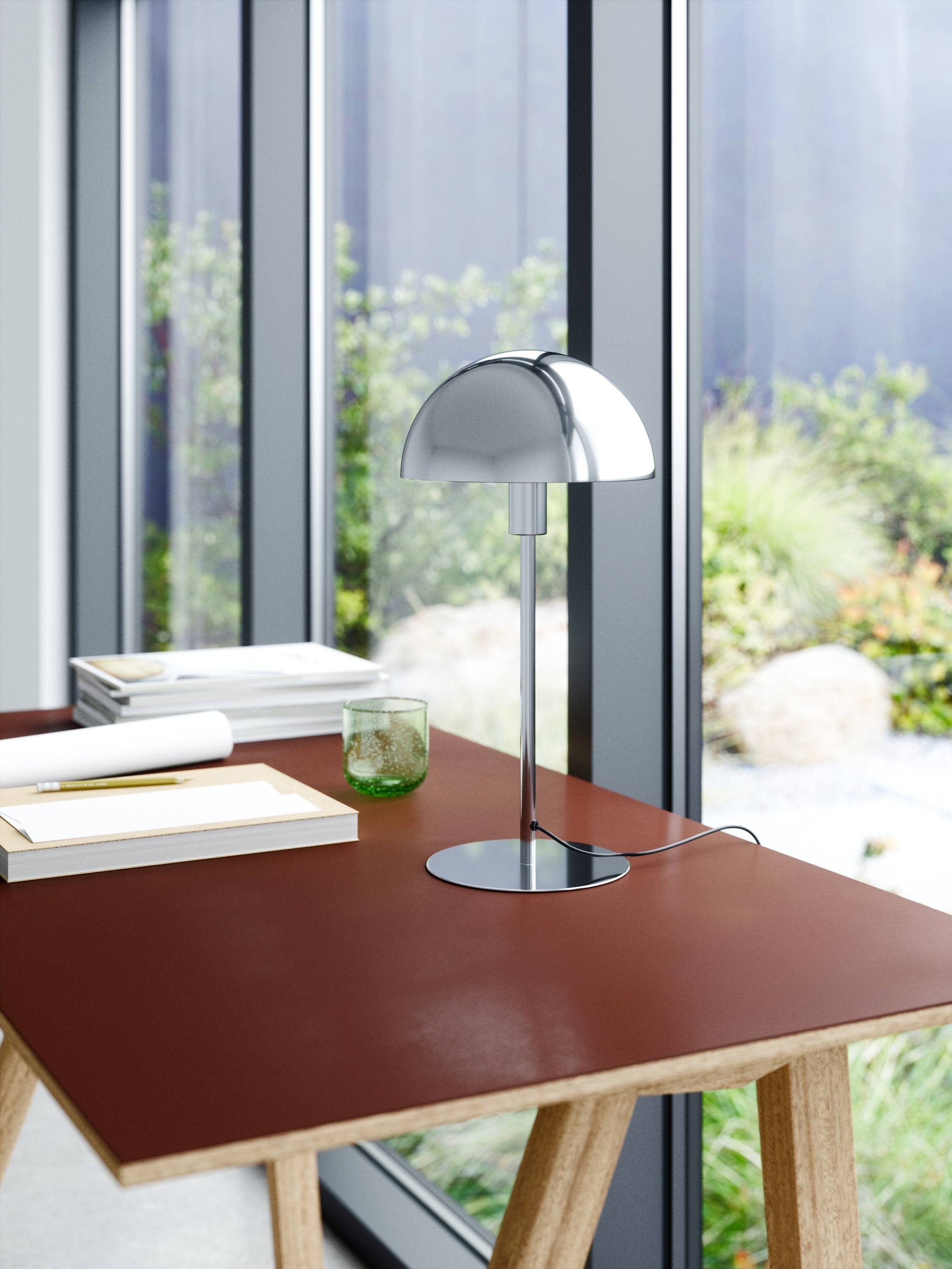 Ein moderner Tisch mit rostroter Oberfläche, darauf eine silberne Lampe und Bücher. Im Hintergrund große Fenster mit Gartenblick.