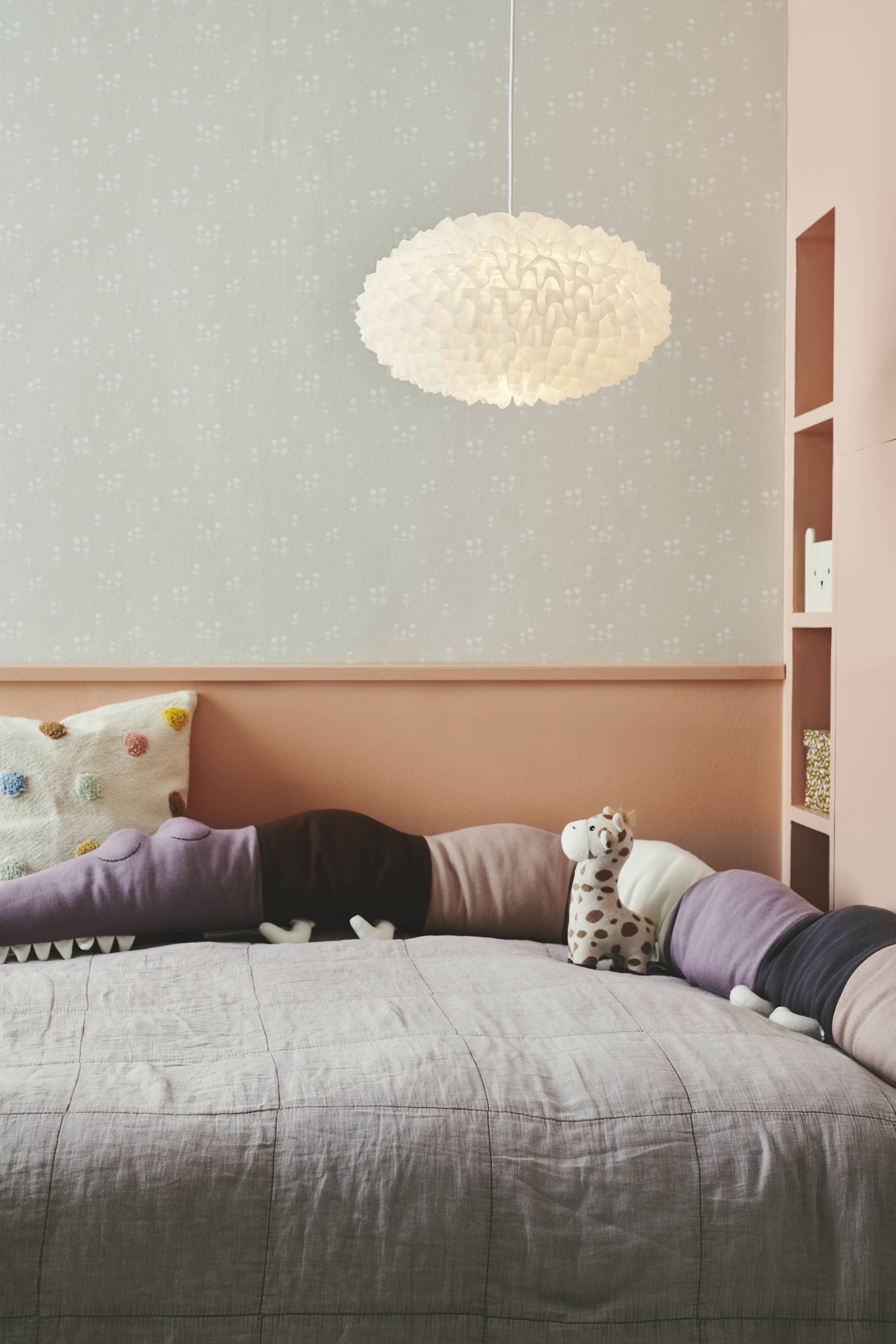 Ein Kinderzimmer mit einem Bett, dekoriert mit bunten Kissen und Stofftieren. Eine interessante Pendelleuchte hängt von der Decke.