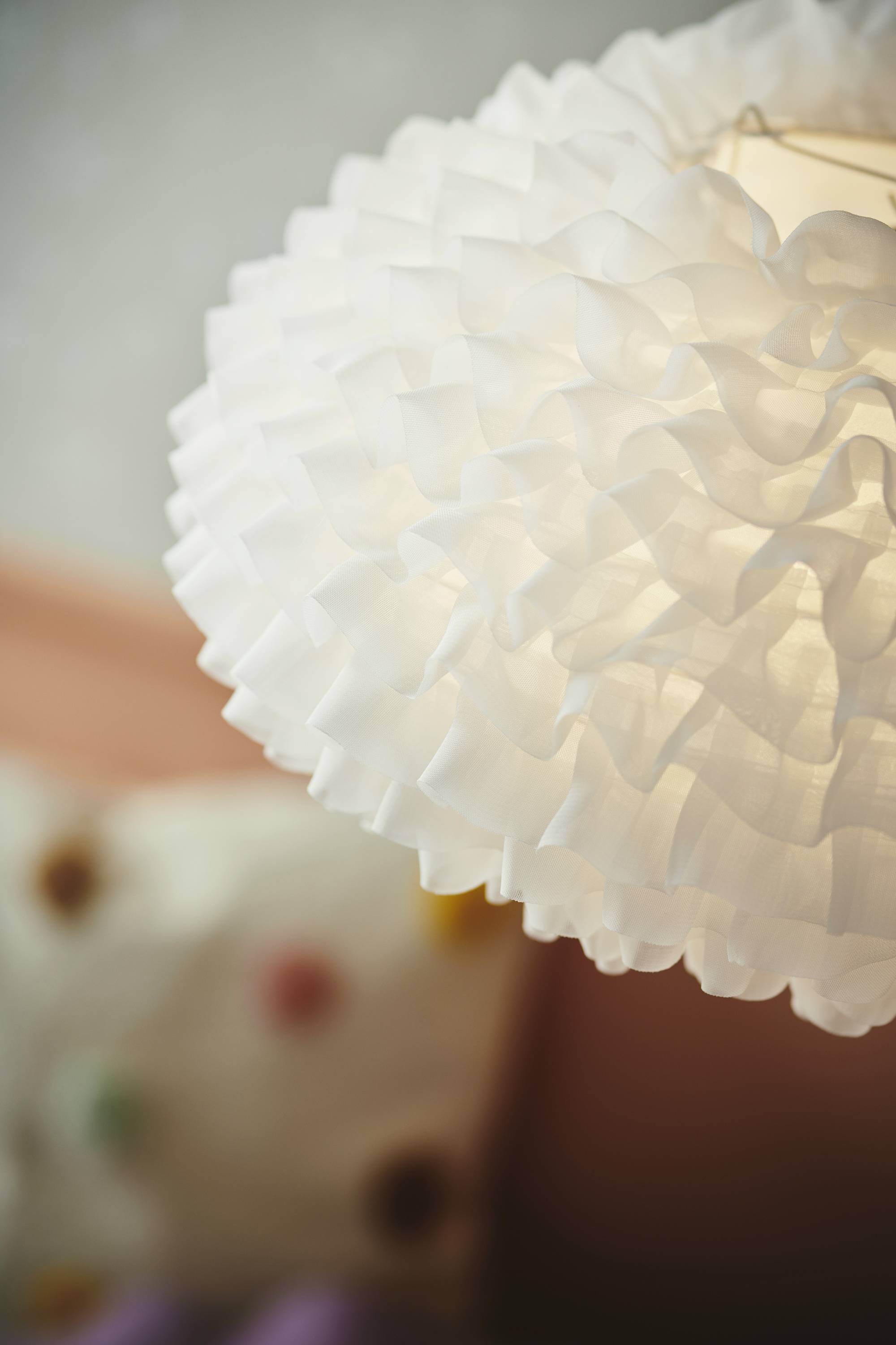 Weiße, runde Deckenlampe mit welliger Struktur hängt von der Decke. Im Hintergrund sind verschwommen bunte Kissen zu sehen.