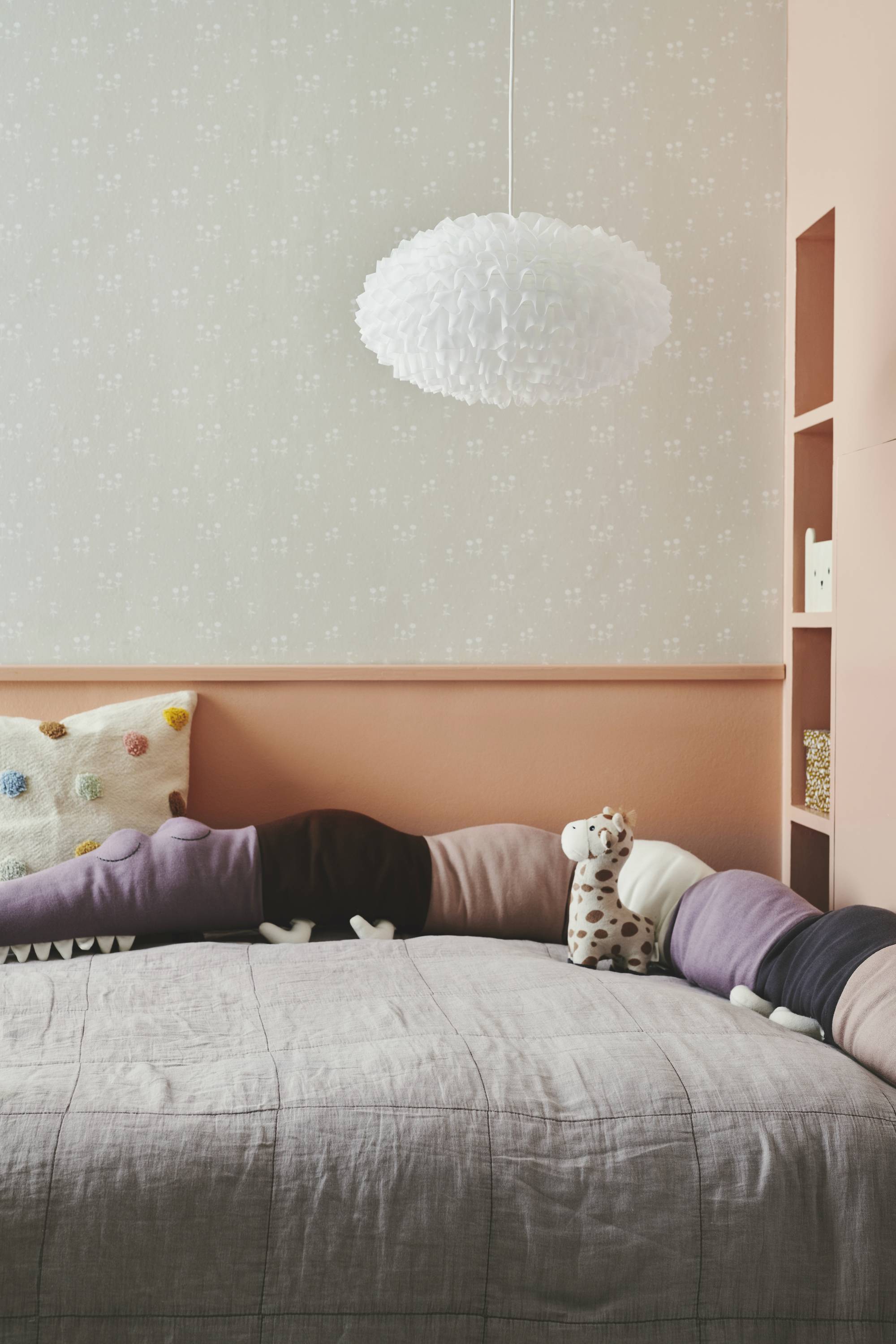 Ein gemütliches Kinderzimmer mit einem Bett, das von bunten Kissen und Plüschtieren umgeben ist. Eine weiße Hängelampe hängt oben.