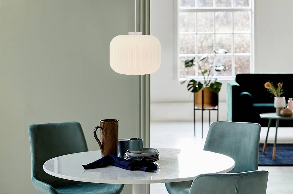 Runder Tisch mit blauer Tischdecke, braune Tasse und weißer Lampe. Im Hintergrund grüne Stühle, Sofa und Topfpflanzen nahe Fenster.