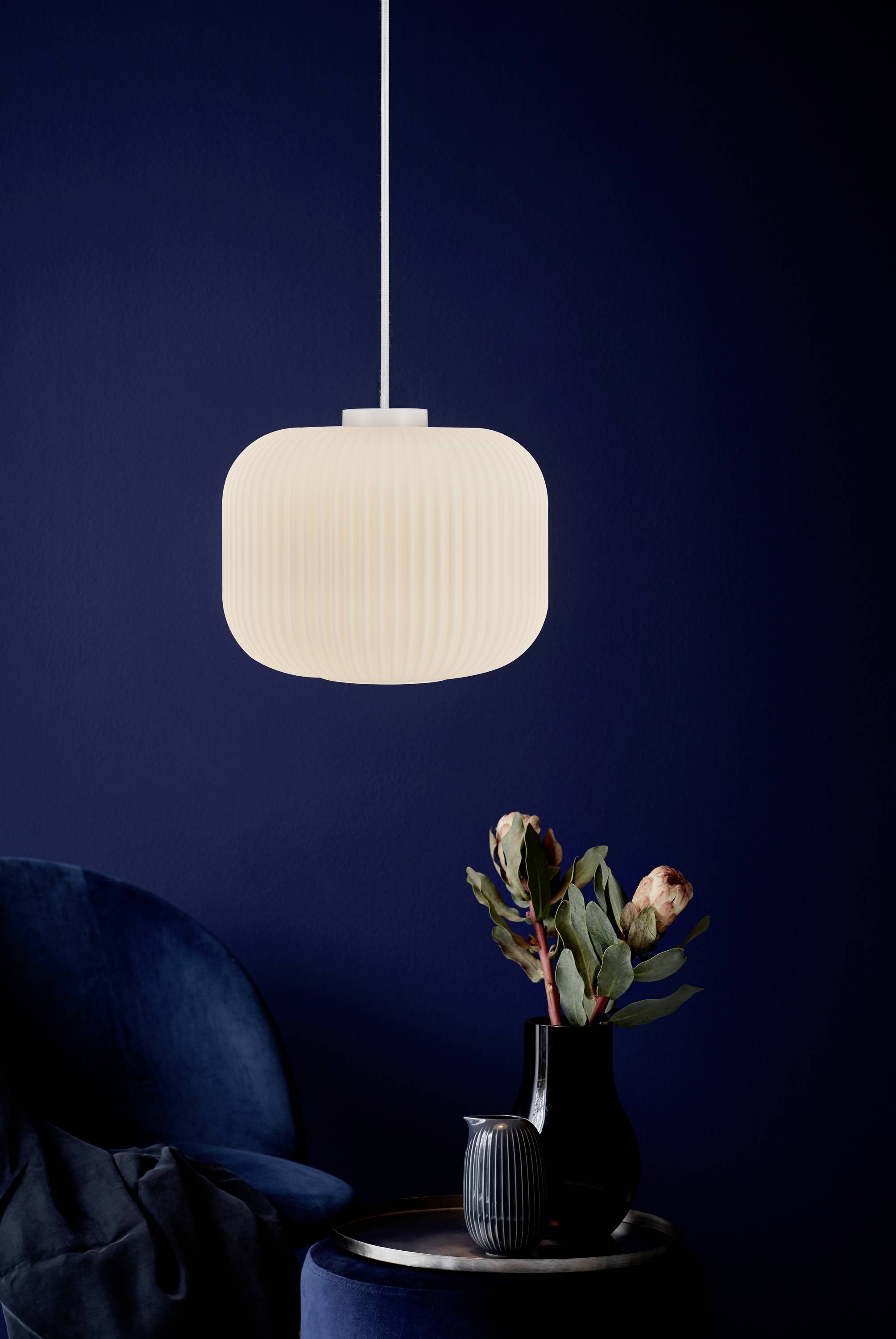 Helle, hängende Lampe über blauem Sessel und Tisch mit dunkler Vase und Blumenarrangement vor dunkelblauem Hintergrund.