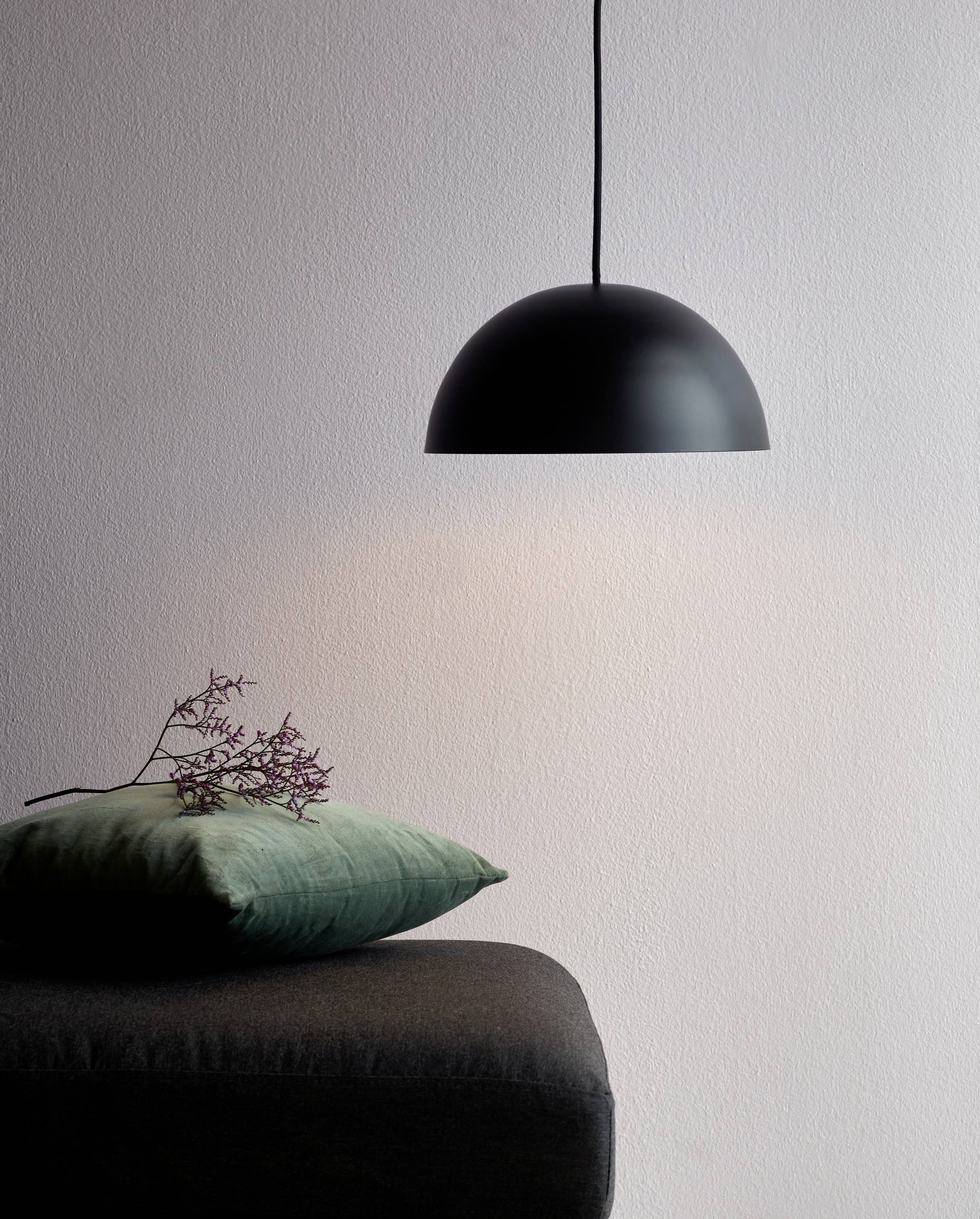 Eine schwarze Pendelleuchte hängt über einem dunkelgrauen Polsterhocker, auf dem ein grünes Kissen mit lila Zweigen liegt.