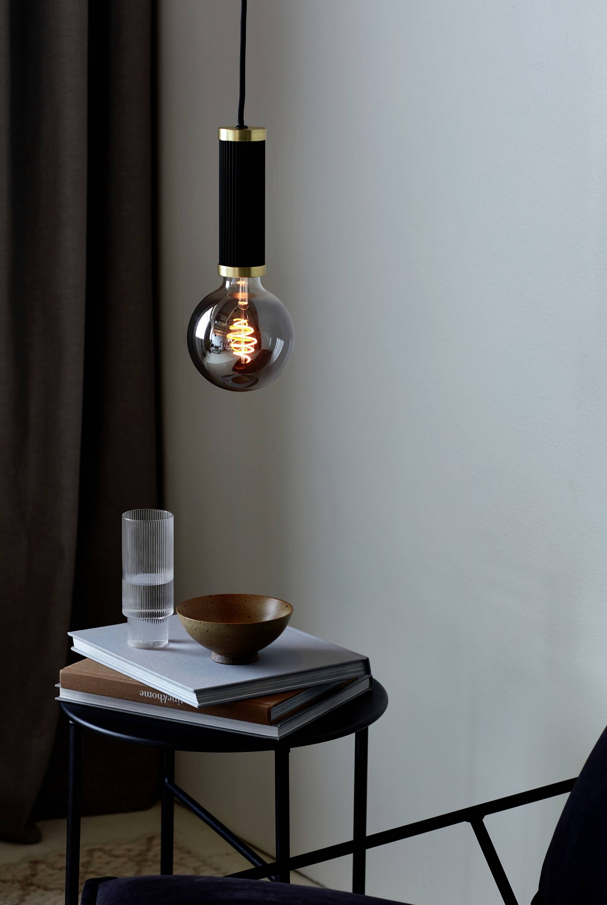 Eine hängende Glühbirne über einem kleinen Tisch mit einem Glas Wasser, einem Holzschälchen und Büchern in einem minimalistisch eingerichteten Raum.