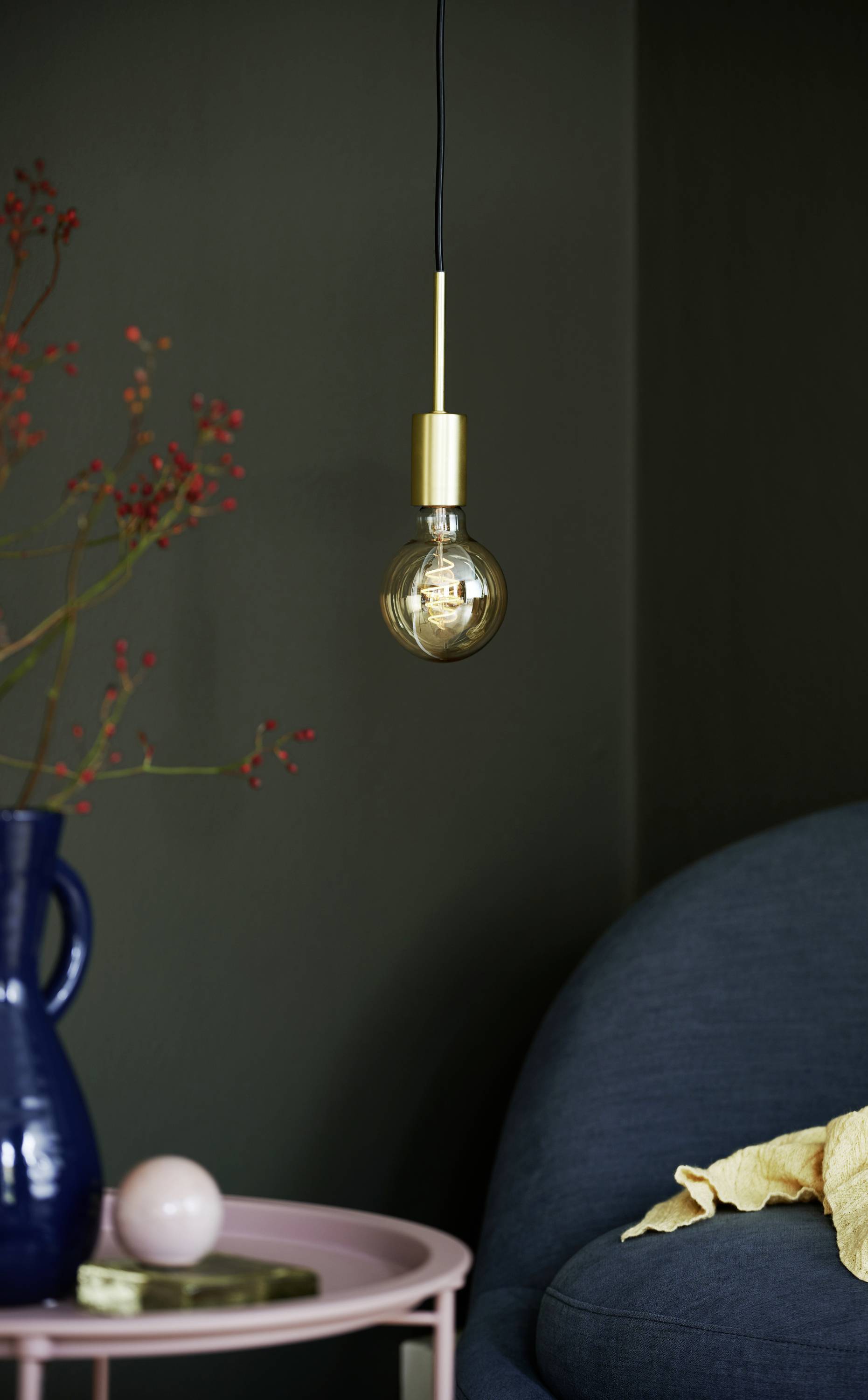 Hängende Glühbirne in modernem Raum, neben blauem Sofa und Tisch mit Vase und Zweigen. Warmes Licht schafft gemütliche Atmosphäre.
