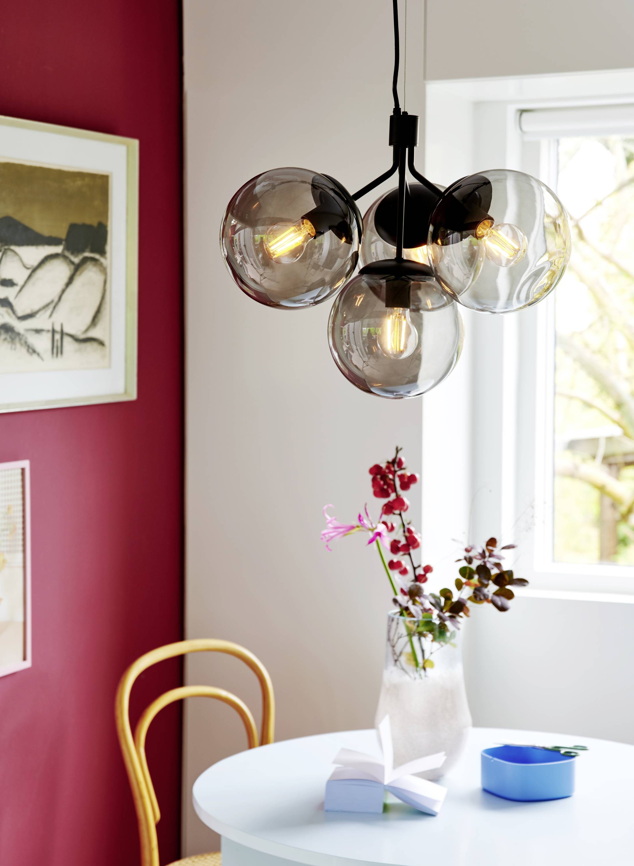 Ein Essbereich mit rundem Tisch, darauf eine Vase mit Blumen. Eine moderne Hängelampe mit vier kugelförmigen Leuchten hängt darüber. Im Hintergrund ein Fenster und ein Wandbild.