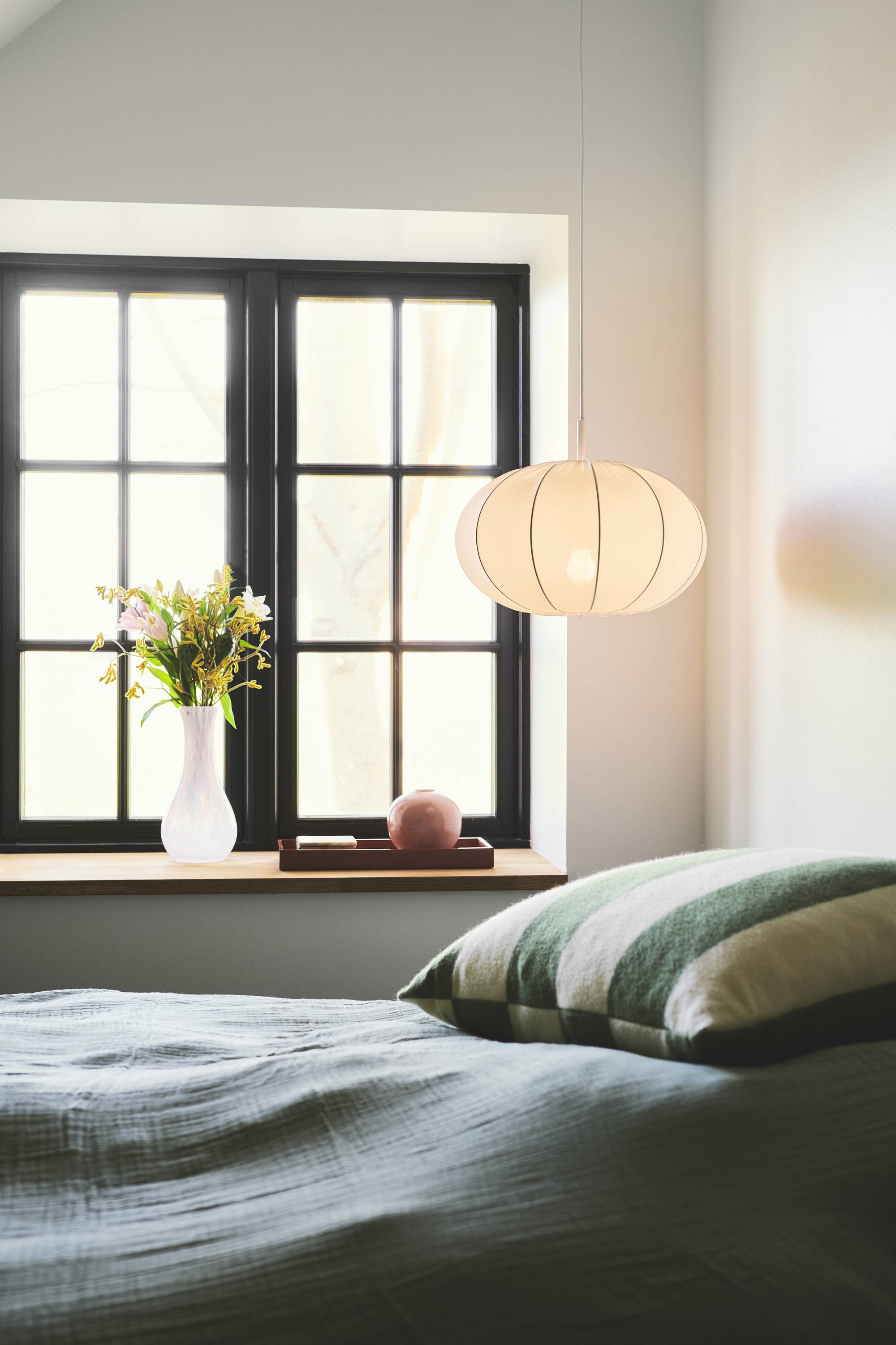 Schlafzimmer mit einer hängenden Lampe, einer Vase mit Blumen auf der Fensterbank und einem Bett mit grün-gestreiften Kissen.