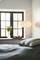 Schlafzimmer mit einer hängenden Lampe, einer Vase mit Blumen auf der Fensterbank und einem Bett mit grün-gestreiften Kissen.