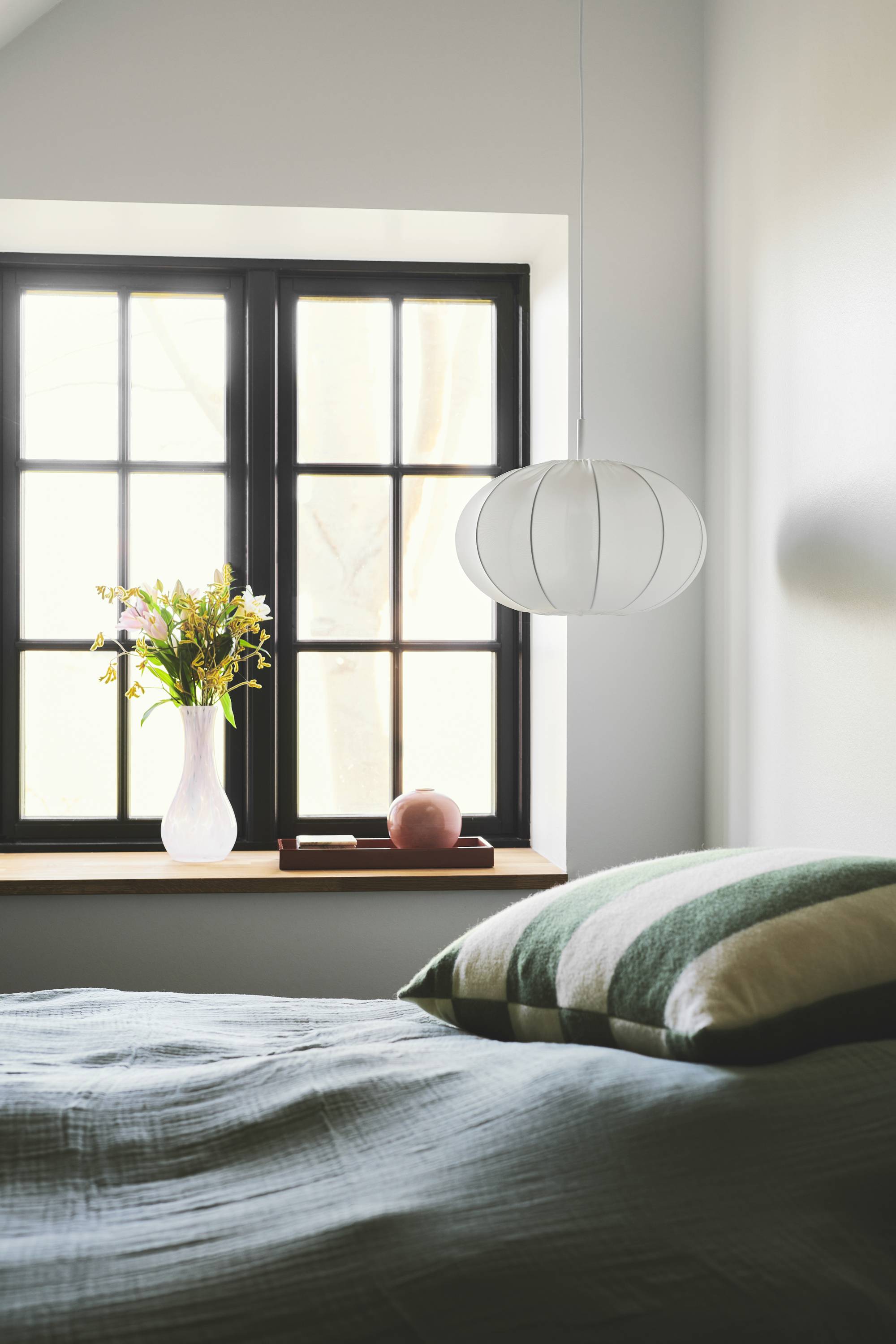 Ein Schlafzimmer mit einem Bett, das große Kissen hat, steht vor einem Fenster. Eine Vase mit Blumen und ein Apfel sind auf der Fensterbank.