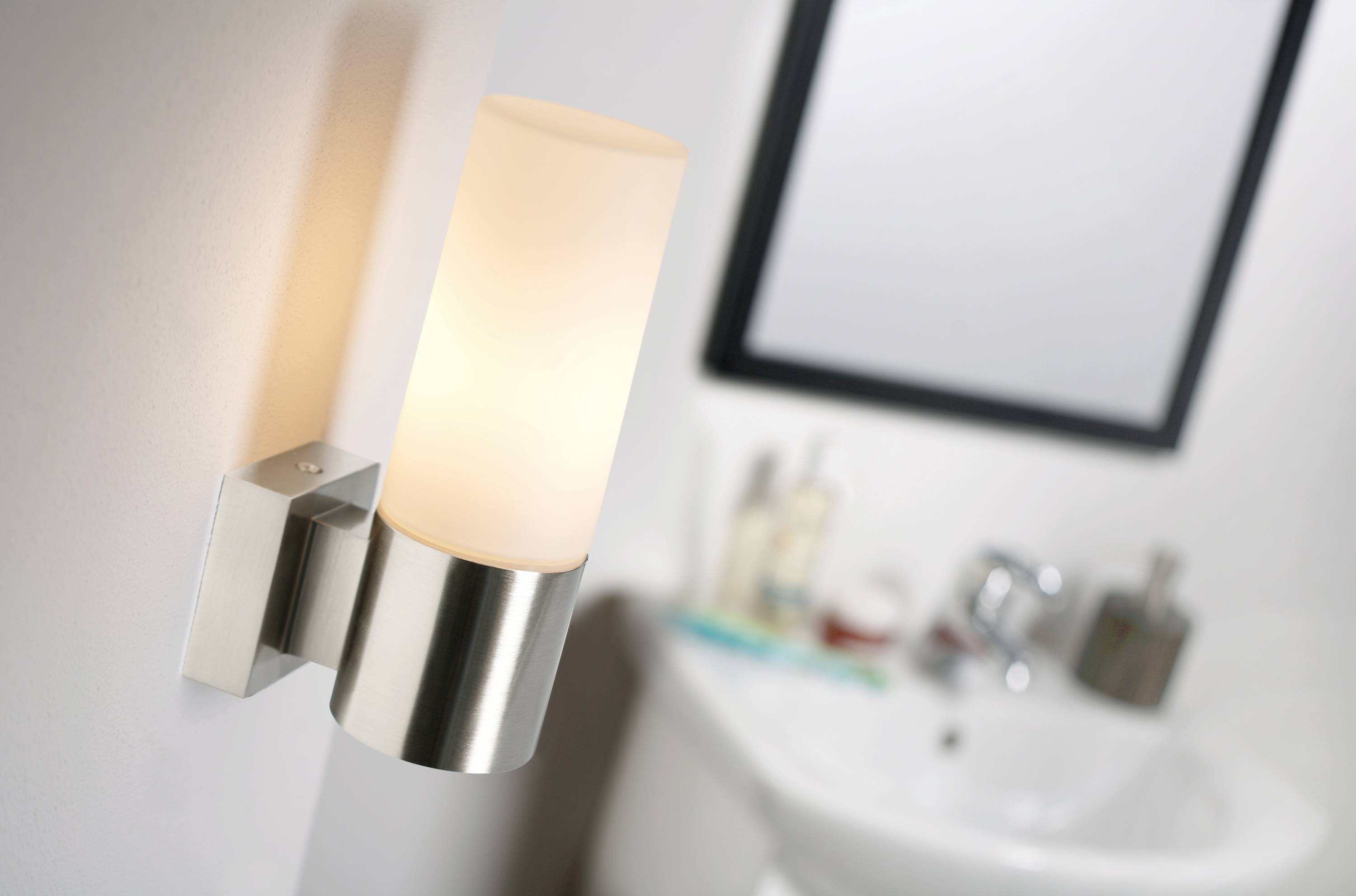 Eine Wandleuchte mit zylindrischem Lampenschirm leuchtet in einem modernen Badezimmer, im Hintergrund sind Waschbecken und Spiegel sichtbar.