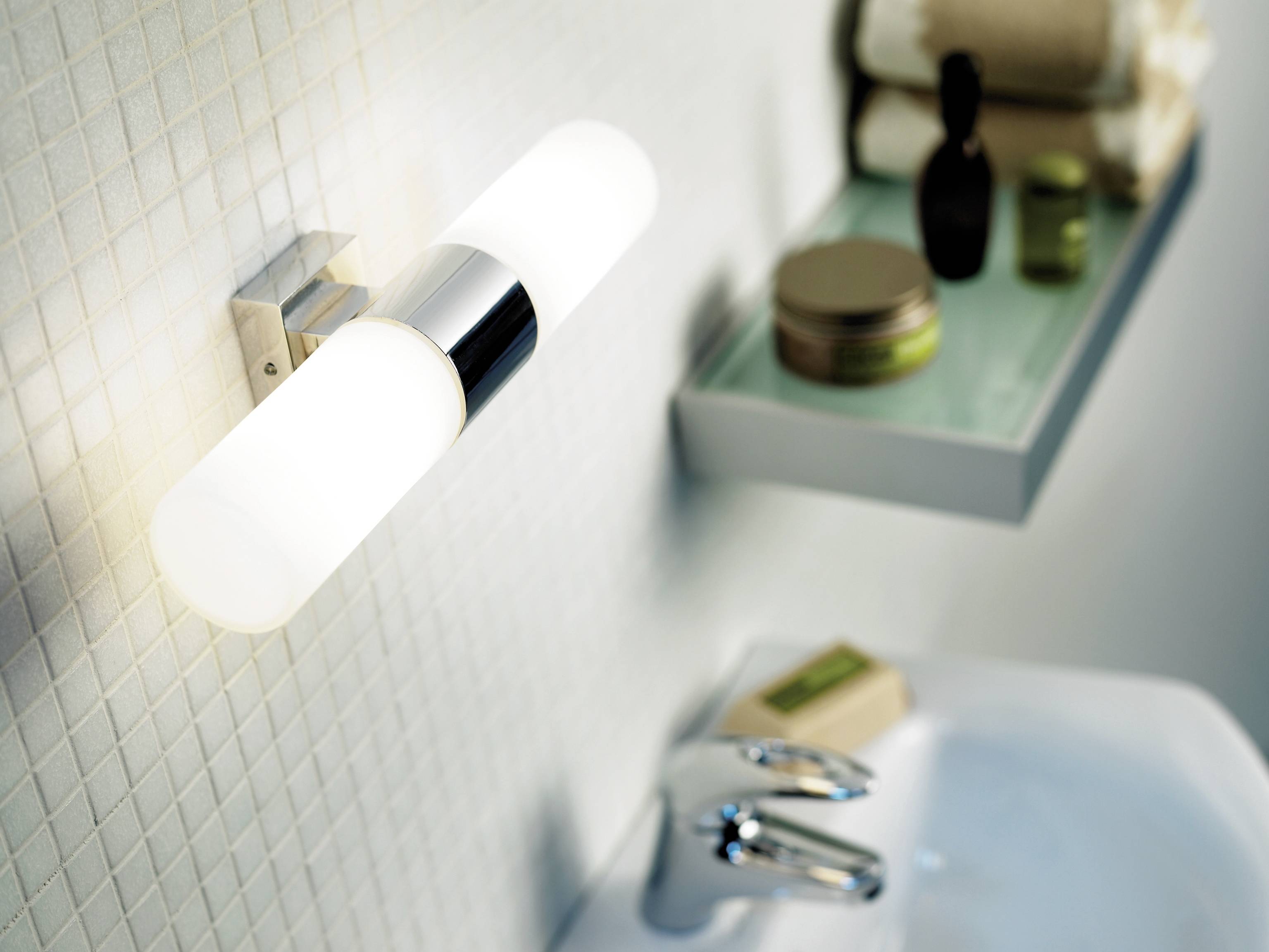 Wandleuchte neben einem Badezimmerwaschbecken mit Glasablage. Auf der Ablage stehen Kosmetikprodukte und Handtücher.