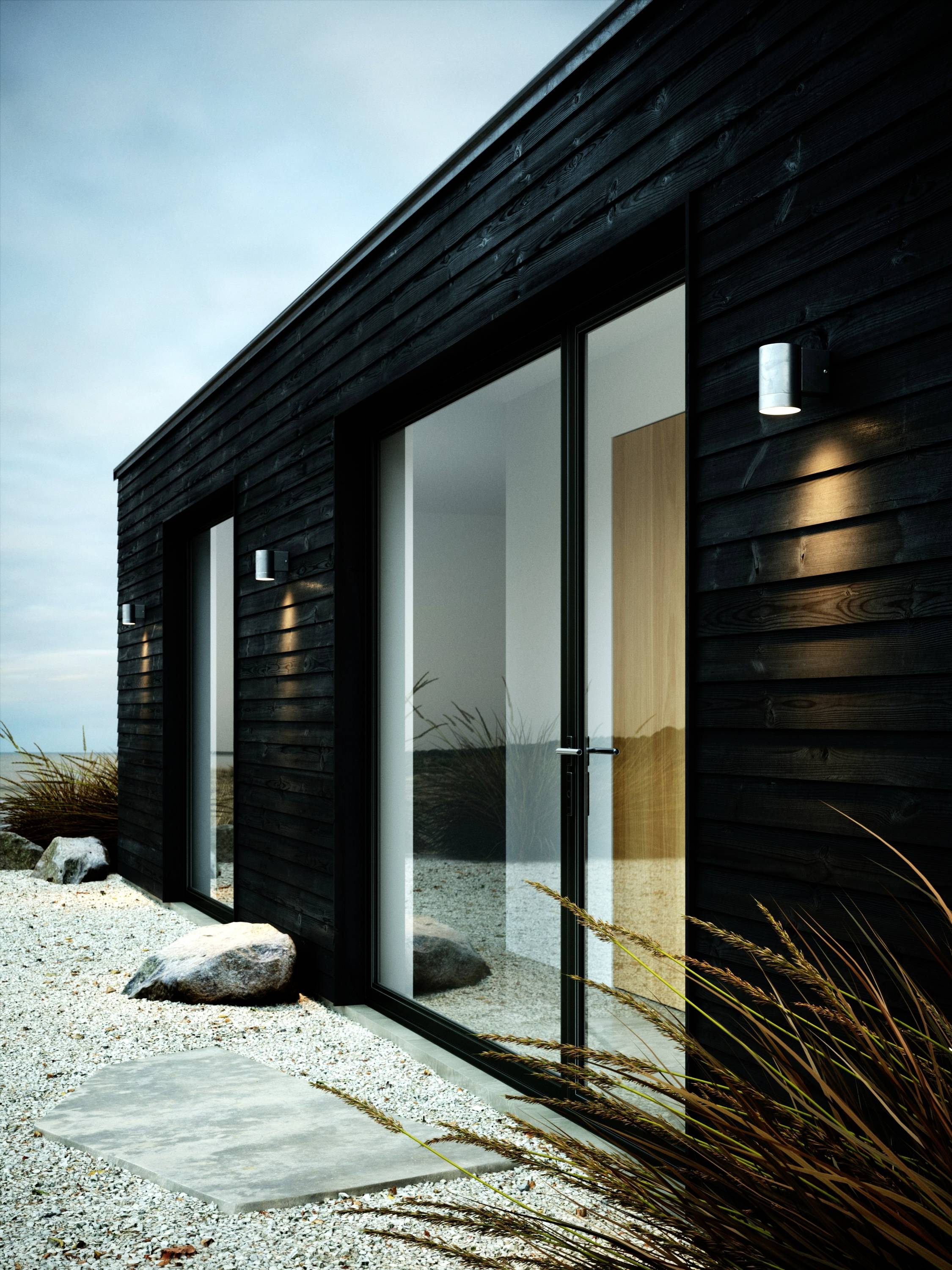 Ein modernes, schwarzes Holzhaus mit großen Glasfronten. Im Vordergrund sind Kies und Gras zu sehen, die eine natürliche Umgebung schaffen.