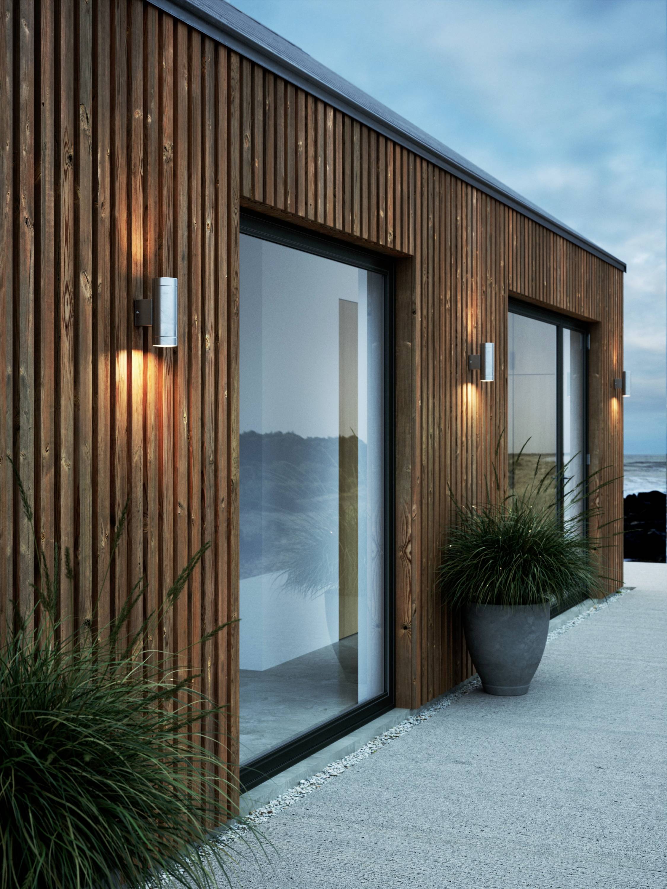 Ein modernes Haus mit Holzverkleidung und großen Glasfronten, beleuchtet von Wandlampen. Daneben stehen Pflanzen in Töpfen auf einem Weg.