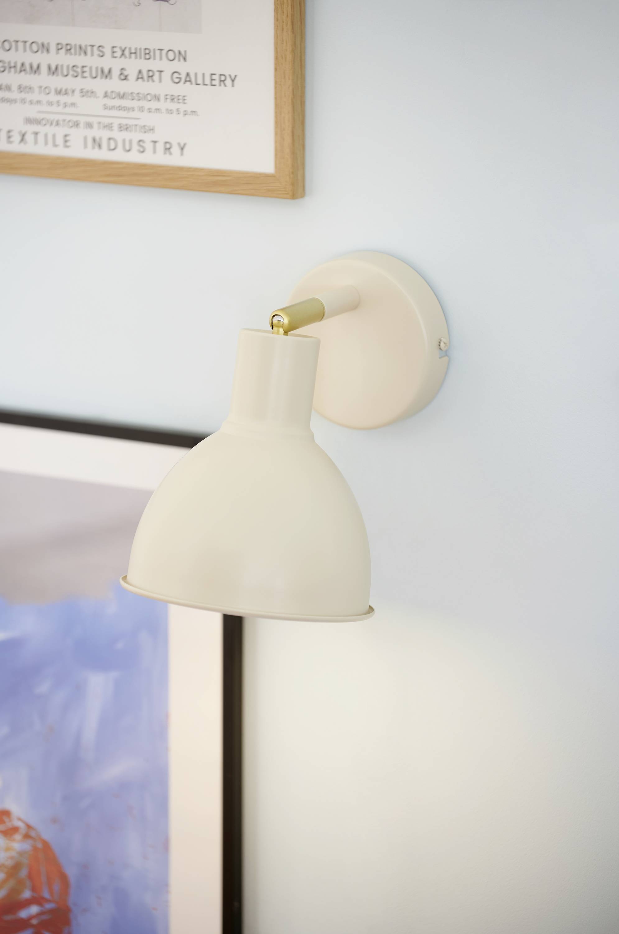 Weiße Wandlampe an blauer Wand, neben gerahmtem Kunstwerk. Oben hängt ein Ausstellungsplakat im Holzrahmen.