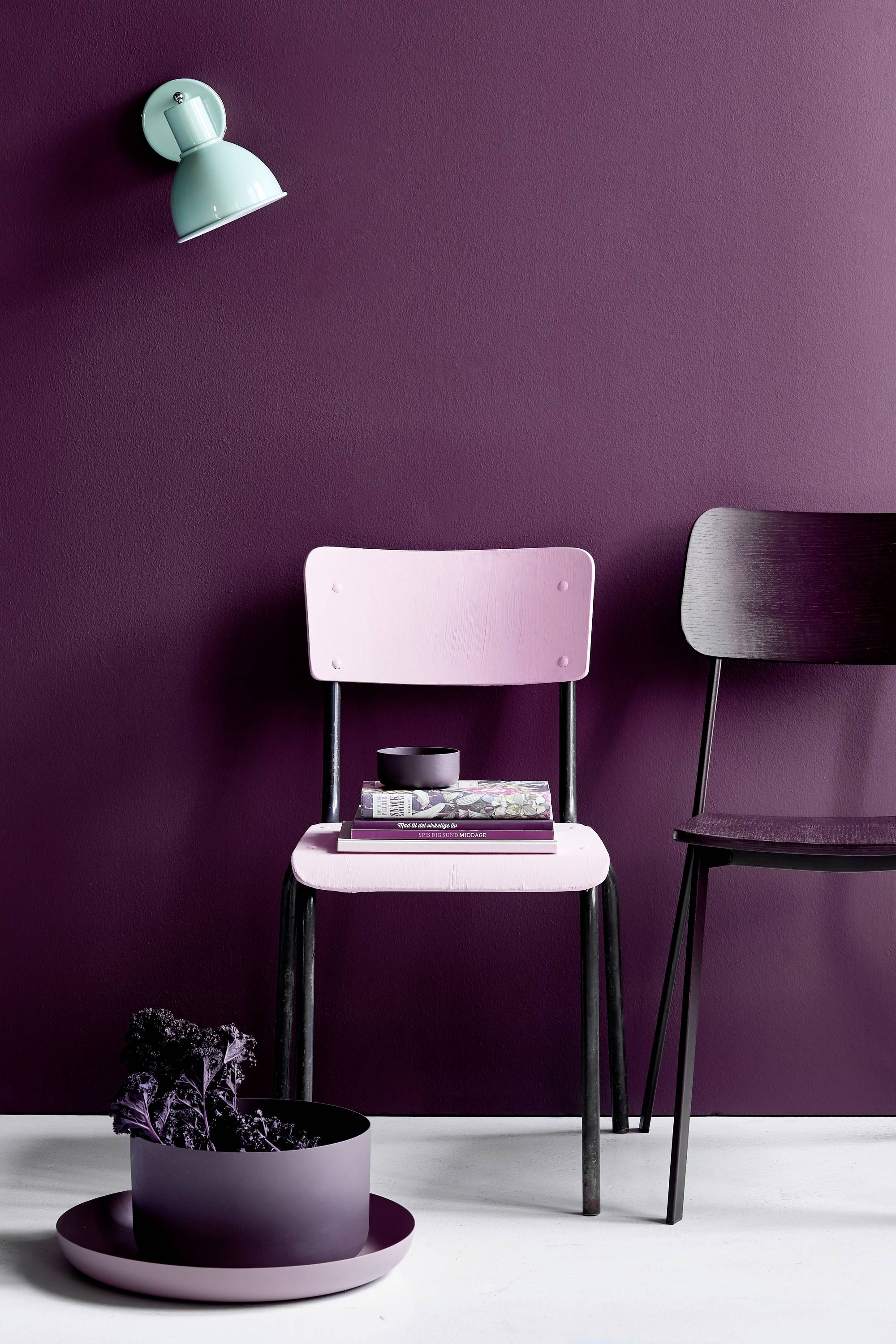 Zwei Stühle vor einer dunkelroten Wand; ein rosa und ein schwarzer Stuhl. Eine grüne Lampe leuchtet auf die Szene.