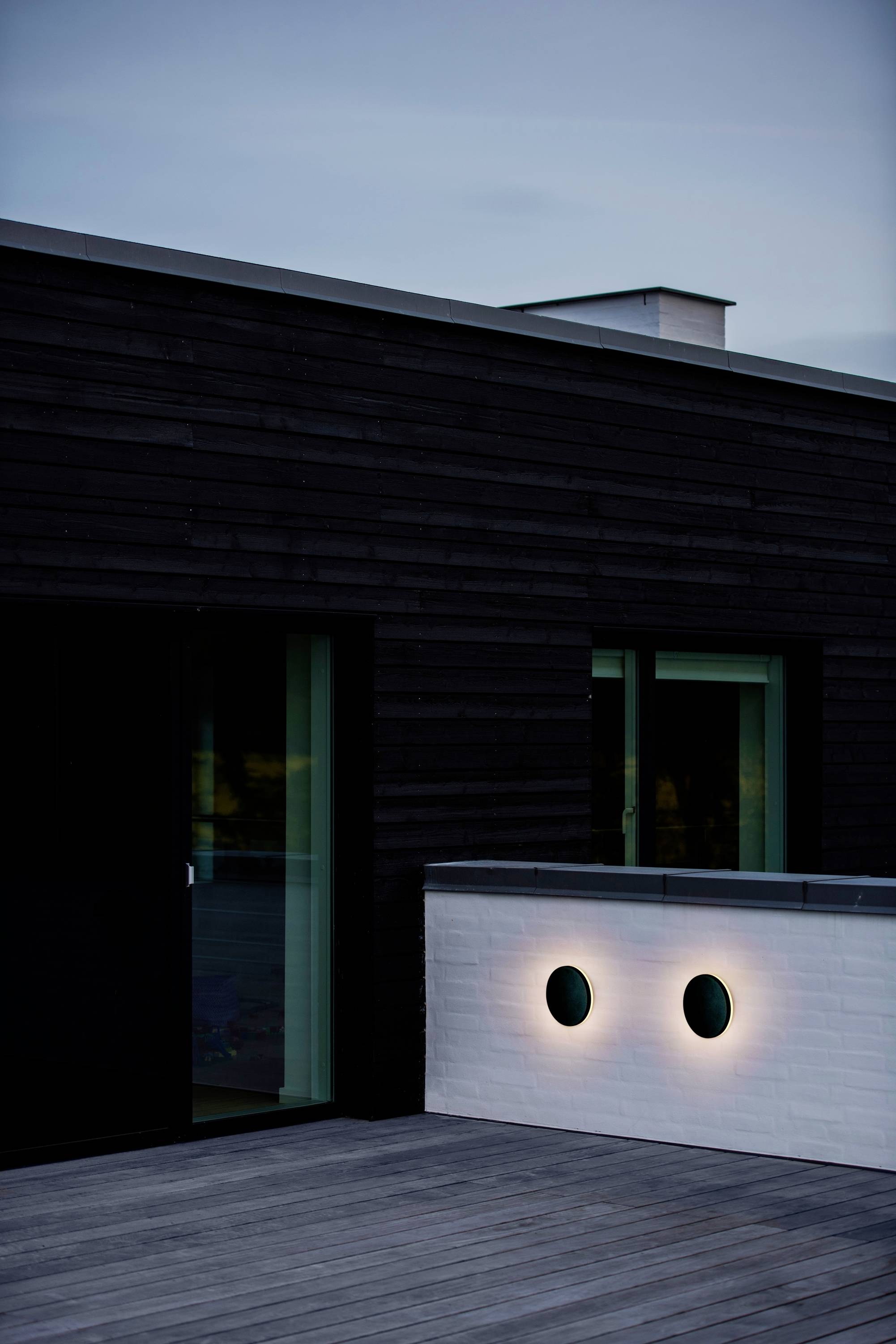 Modernes Haus mit dunklem Holz verkleidet. Zwei runde Lichter neben einer Glastür auf einer weißen Wand sind beleuchtet.