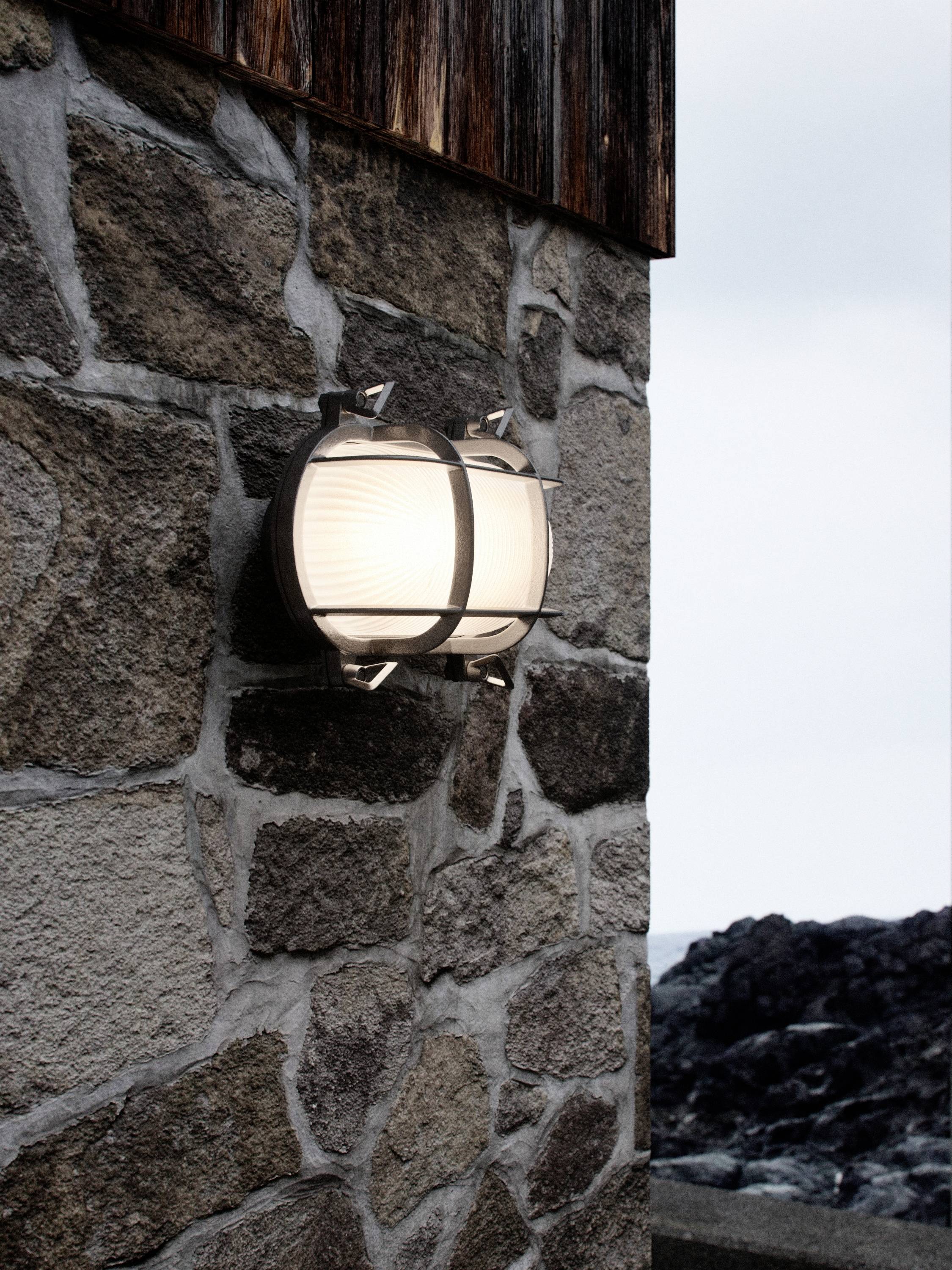 Außenlampe an einer Steinwand, leuchtet in der Dämmerung, neben einer felsigen Küste.